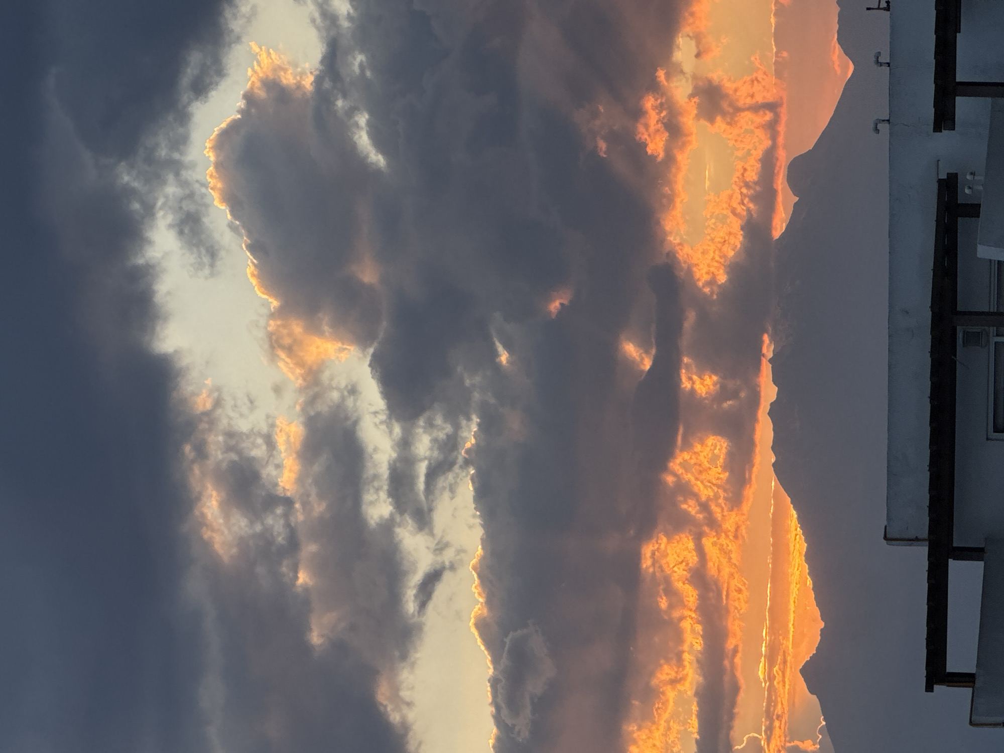 The image depicts a striking sunset in San Francisco Ocotlán, Mexico. Dark, heavy clouds dominate the left side of the frame, contrasting sharply with the bright orange and yellow light illuminating the right side. This warm light seems to emanate from behind a mountain range visible in the distance. A building with a window and some pipes is visible in the upper right corner of the image. The photograph is rotated 90 degrees counter-clockwise.