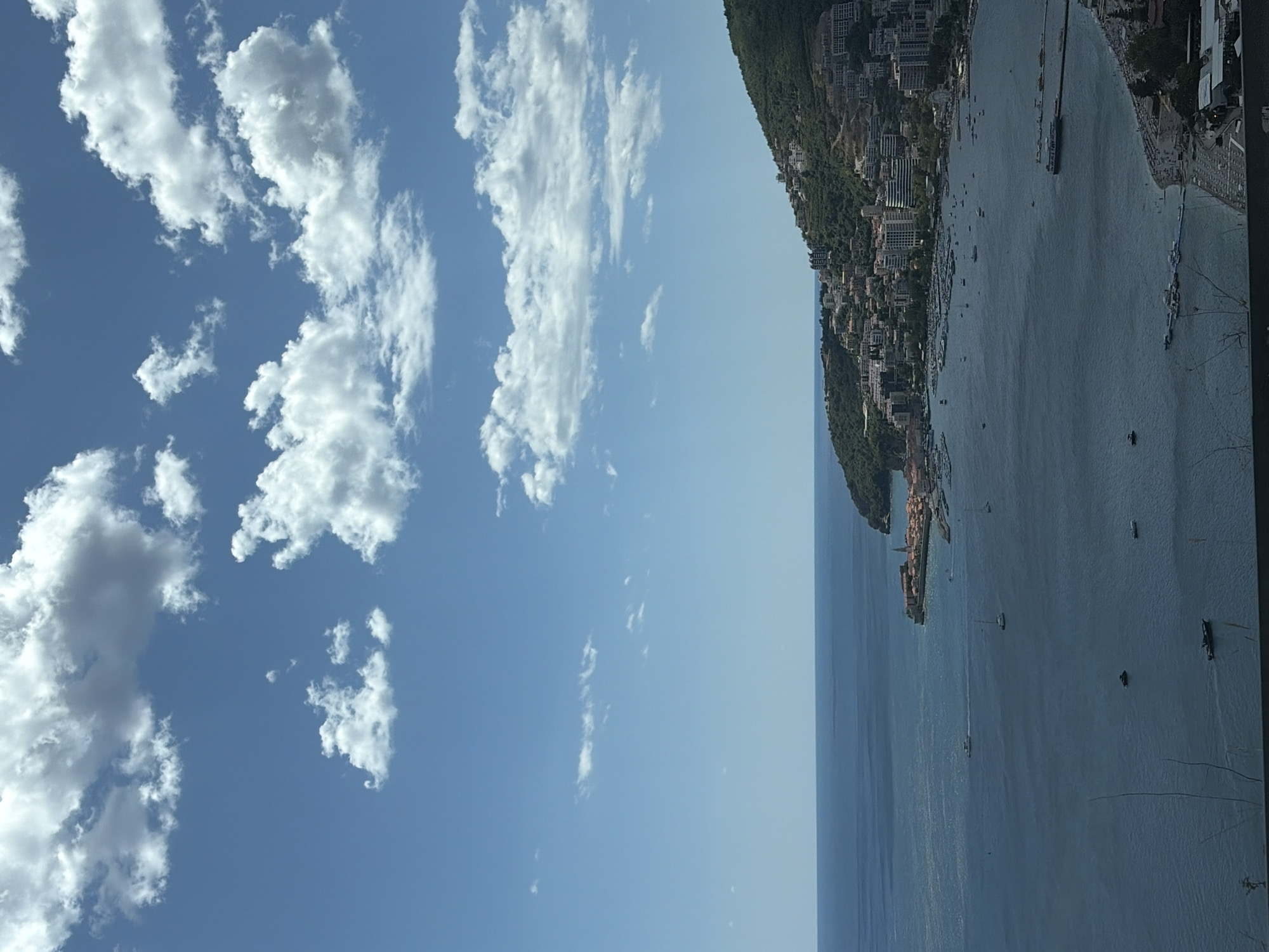 The image captures a panoramic view of Budva, Montenegro, on a sunny day with scattered clouds. The scene is dominated by the expansive blue Adriatic Sea meeting the clear blue sky. In the foreground, a dark silhouette of foliage runs along the bottom edge of the frame. The middle ground features a bustling bay filled with numerous boats, including sailboats and motorboats. A prominent breakwater with several vessels moored alongside extends into the water. Along the shoreline, a wide beach is visible, lined with rows of sun loungers and umbrellas, with a scattering of people enjoying the seaside.
The coastline curves into the background, revealing the old town of Budva with its characteristic red-tiled roofs, clustered around a peninsula. Adjacent to the old town, modern buildings and hotels ascend the lush green hillside that overlooks the bay. The densely built-up slopes are dotted with white and cream structures, indicating a thriving resort town. The overall impression is one of a vibrant coastal city enjoying a beautiful summer day.
