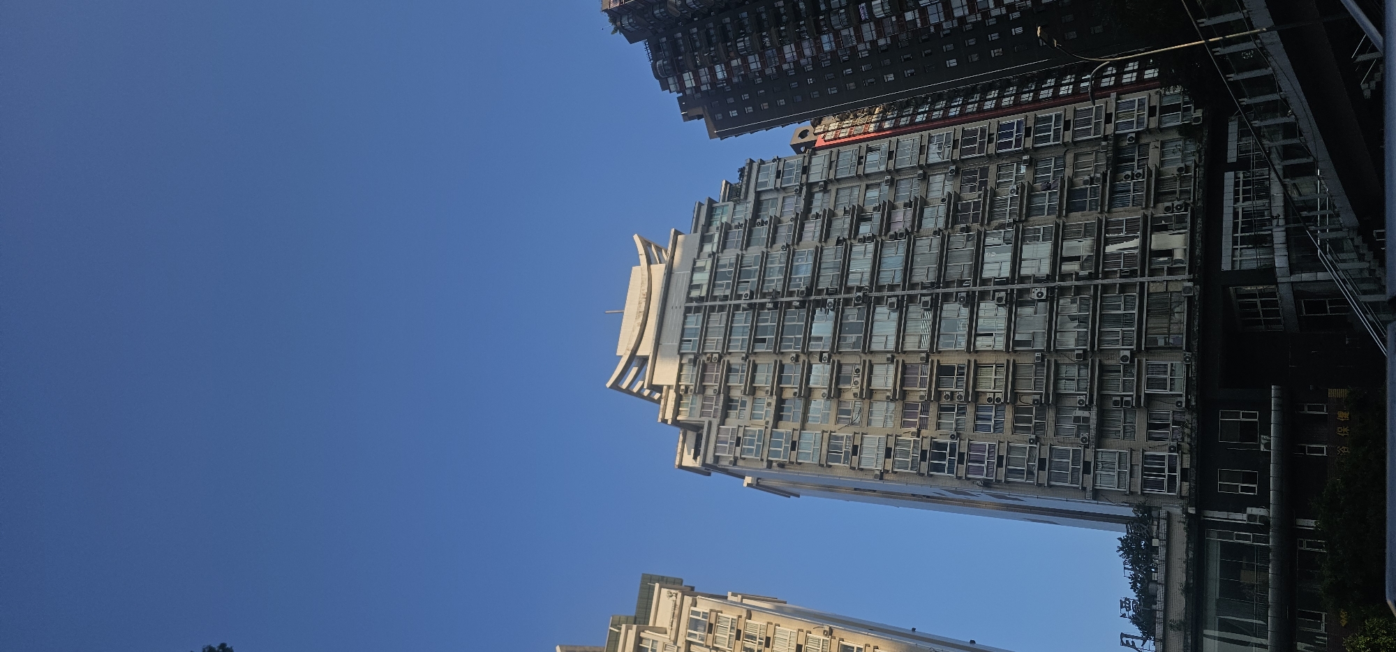 This media file captures a low-angle view of several tall buildings against a clear, bright blue sky, suggesting a sunny daytime setting. The buildings are modern high-rises, densely packed with windows and featuring numerous air conditioning units visible on their facades. The perspective emphasizes the height and scale of the architecture.
The central building is particularly striking, with a unique, layered design that steps inwards as it ascends, topped by a curved, light-colored element. To the upper left, another building is visible, appearing darker and perhaps older, with a more uniform facade. At the bottom, a portion of another building is seen, also constructed with many windows.
The scene is urban, likely a densely populated city. The image doesn't show any people, focusing entirely on the built environment. The sunlight casts some shadows and highlights the textures of the building materials.
Visible text can be seen on one of the buildings on the right side. Some characters are discernible, appearing to be Chinese. One sign seems to read "浴保健" which translates to "bath and health care," and another sign is partially visible with characters like "福" (fortune) and "緣" (fate).
The overall impression is one of a bustling, contemporary urban landscape under a serene sky.