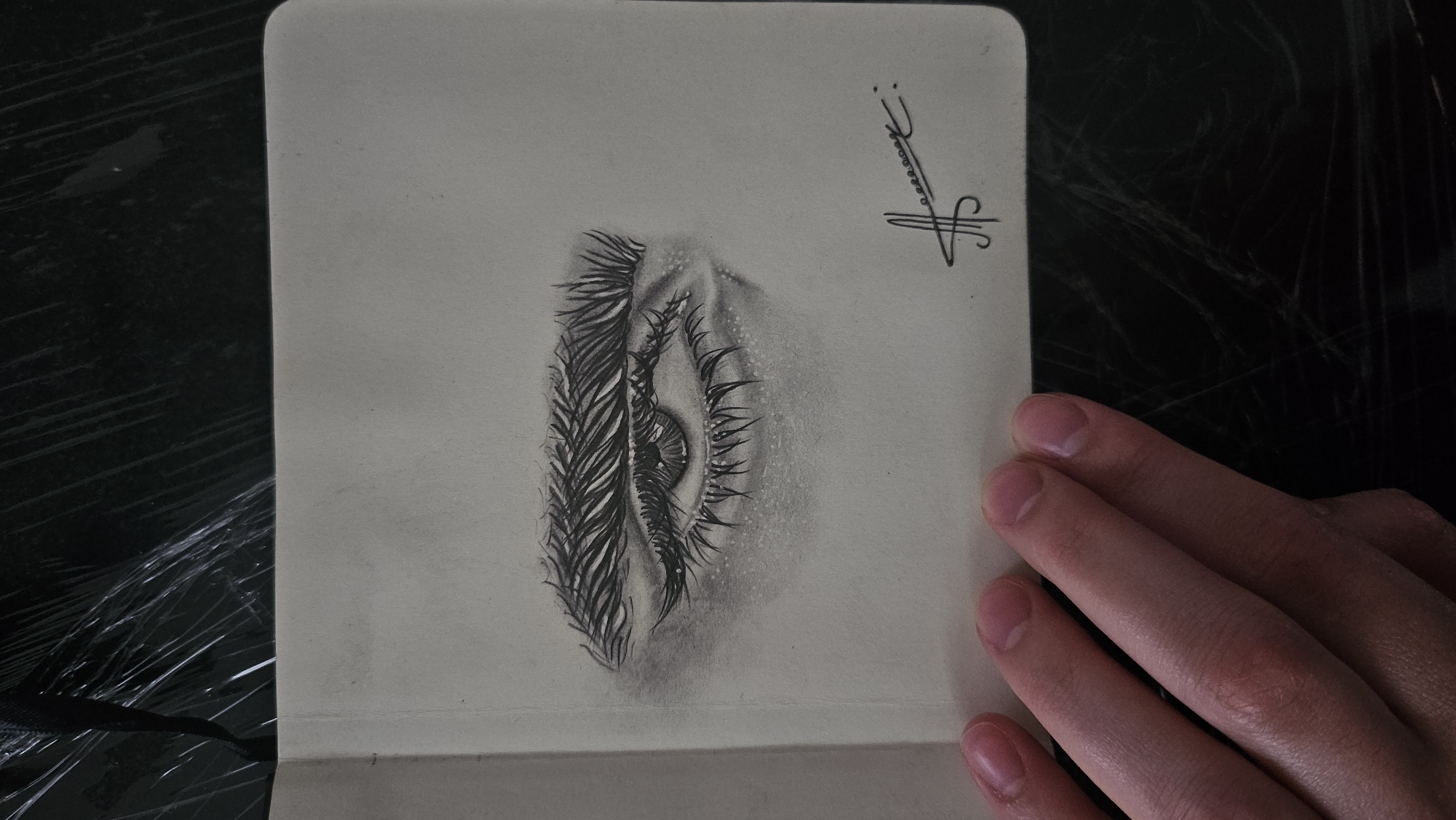 A grayscale pencil sketch of a human eye and eyebrow is shown on a tan-colored page within a sketchbook. The eye is rendered with detailed shading, capturing the texture of the eyelashes, the iris of the eye, and subtle highlights. The eyebrow above is also depicted with individual strokes. To the lower right of the sketch, a stylized signature in black ink is present. A person's hand with visible fingernails rests on the bottom left portion of the page, partially obscuring the sketch. The background of the image is dark and appears to be a textured surface, possibly covered in plastic wrap. The scene is set in an indoor environment, with no discernible city or location details.