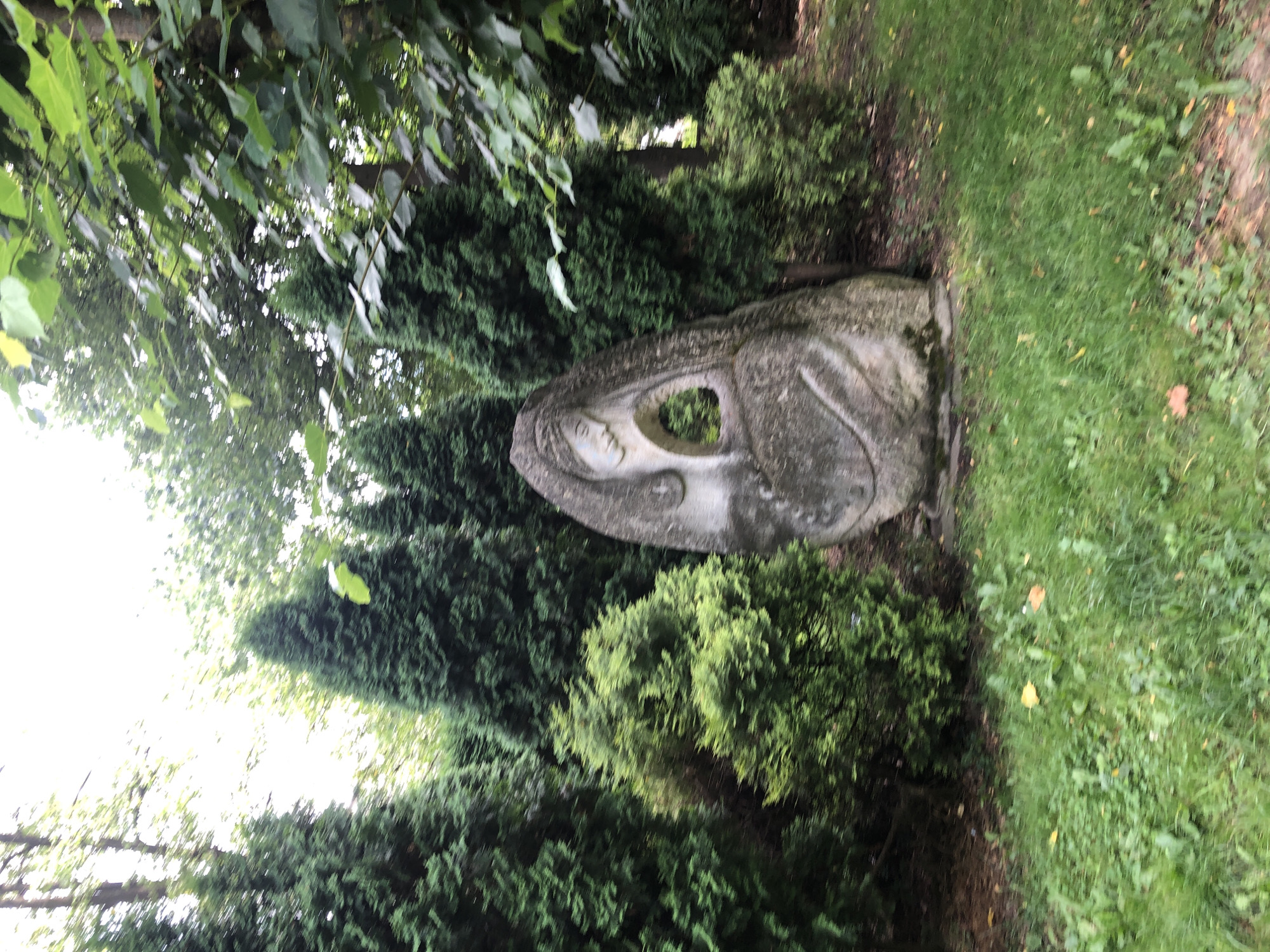 A stone sculpture of a woman's head and upper torso is situated in a park-like setting, surrounded by various evergreen trees and shrubs. The sculpture is rough-hewn, with carved features depicting a profile of a woman with her head tilted downwards and her eyes closed. A circular opening is present in the center of the sculpture. The sculpture is positioned on a patch of grass, with more trees and foliage visible in the background. The overall scene is naturalistic and serene. The location is the Memorial of Ukrainian Recruits in Kolomyia, Ukraine.