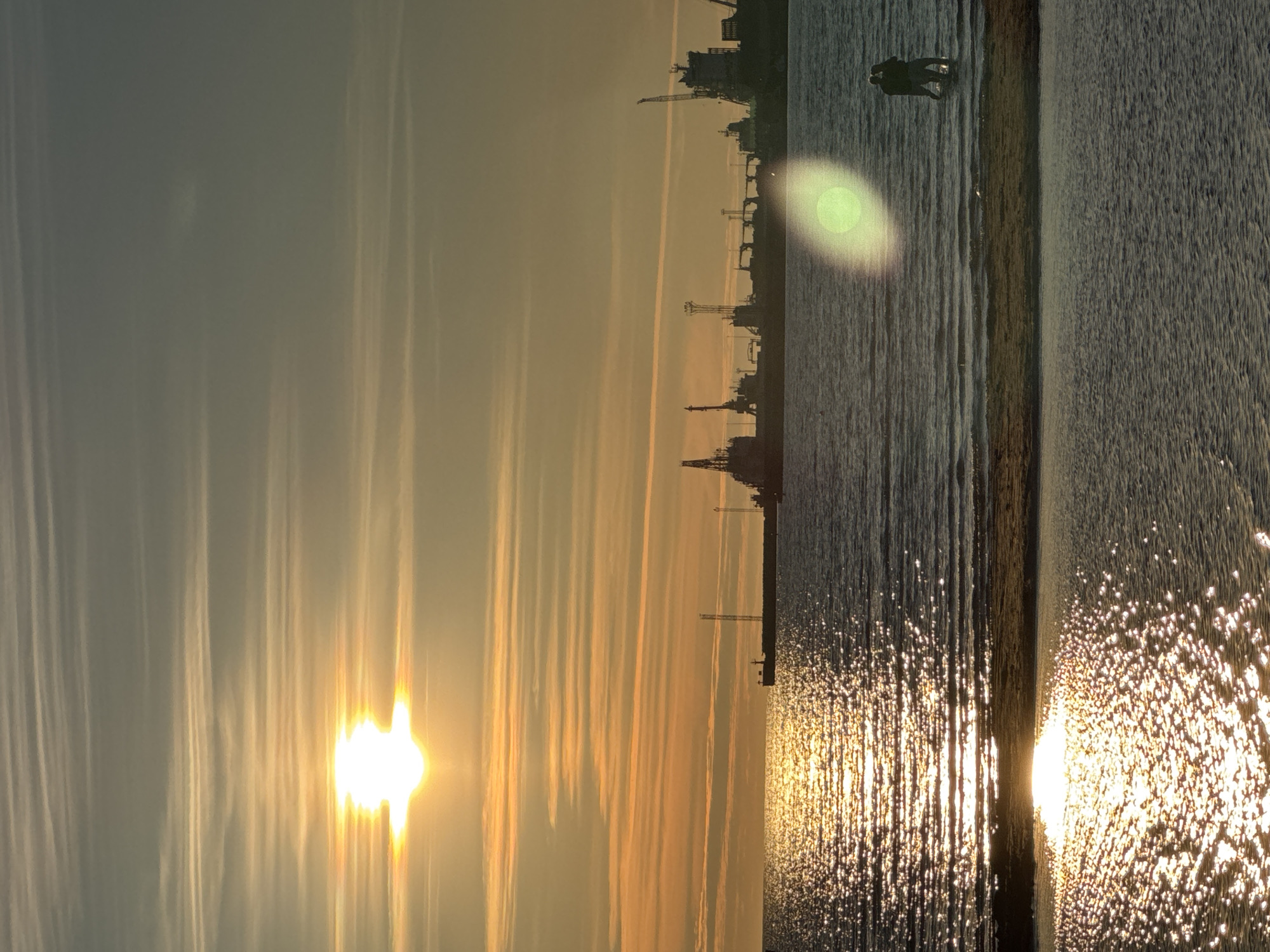 The image depicts a serene sunset over the Baltic Sea, likely from the shores of Tallinn, Estonia, given the presence of the Tallinn Port industrial skyline in the background. The sun, a brilliant orb, casts a warm golden glow across the sky, illuminating the rippling water with shimmering reflections. Delicate, wispy clouds stretch horizontally, adding texture to the sky.
In the mid-ground, silhouetted against the vibrant sunset, is the industrial landscape of the Tallinn Port. Numerous cranes and cargo ships are visible, forming a stark contrast to the natural beauty of the sea and sky.
In the foreground, a couple stands partially submerged in the shallow water, their figures silhouetted. They appear to be embracing or standing close together, creating a sense of intimacy against the grand backdrop of the sunset and the industrial port. The water in the foreground is calmer, with a sandy or pebbled seabed visible beneath the surface in some areas. A subtle lens flare, appearing as a pale green orb, is present in the mid-ground, slightly obscuring the view of the couple and the port. The overall scene evokes a sense of peaceful contemplation, juxtaposing human connection with the vastness of nature and industry.
