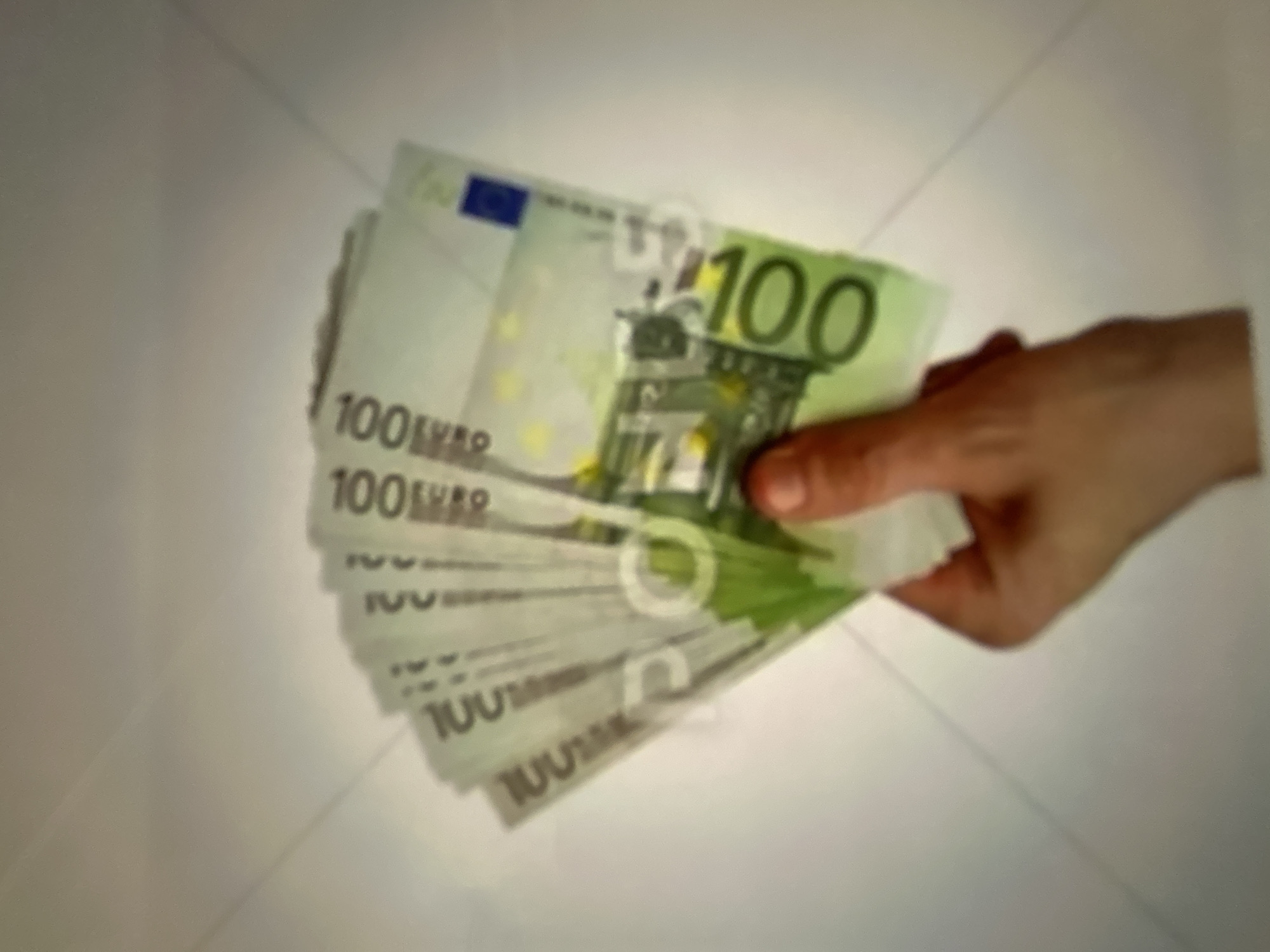 A hand is holding a fan of 100 euro banknotes. The background is a plain, light-colored surface with faint diagonal lines. The image is out of focus. There is no discernible location in Gelendzhik, Russia.