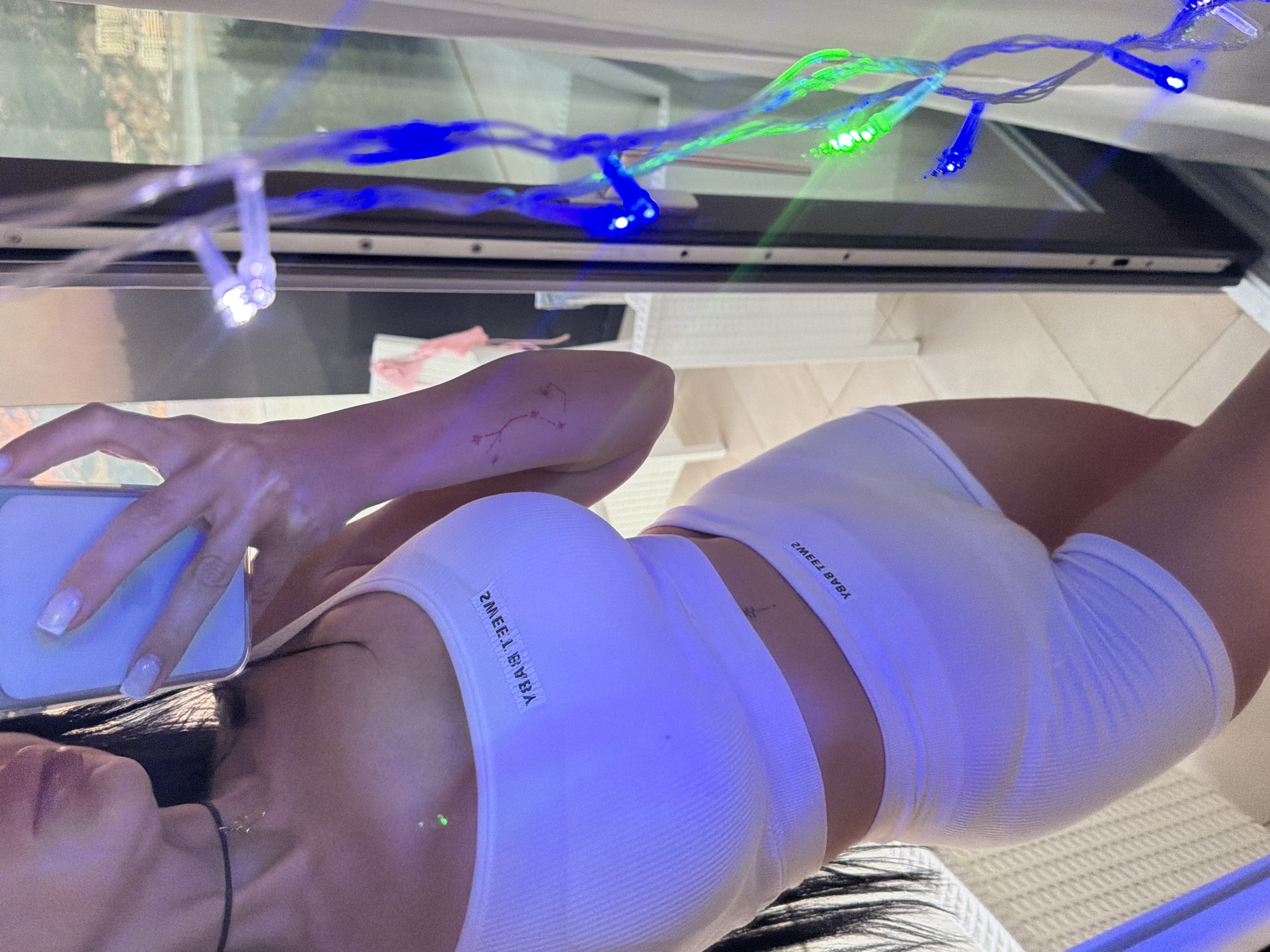 A woman wearing a white crop top and shorts with "SWEET BVBA" printed on them is taking a selfie in an indoor setting. Blue and green string lights are draped overhead. The location is possibly in a residence in Boreti, Montenegro, though this cannot be definitively determined from the image alone.
