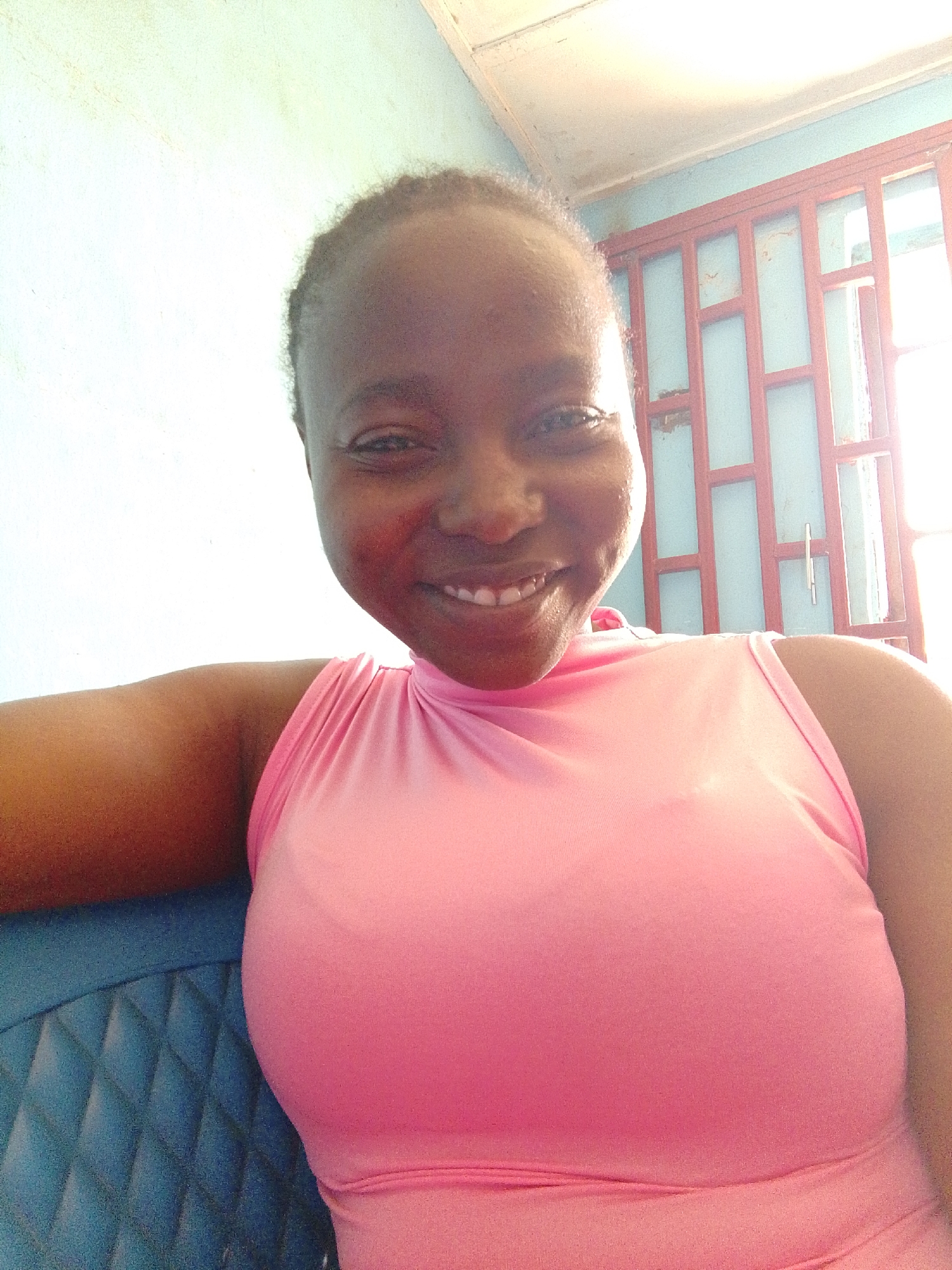 A young woman with dark skin and short, dark hair smiles at the camera. She is wearing a pink sleeveless top and is seated on a blue chair. Behind her is a light blue wall and a window with a red frame. The location is Jos, Nigeria, as specified.