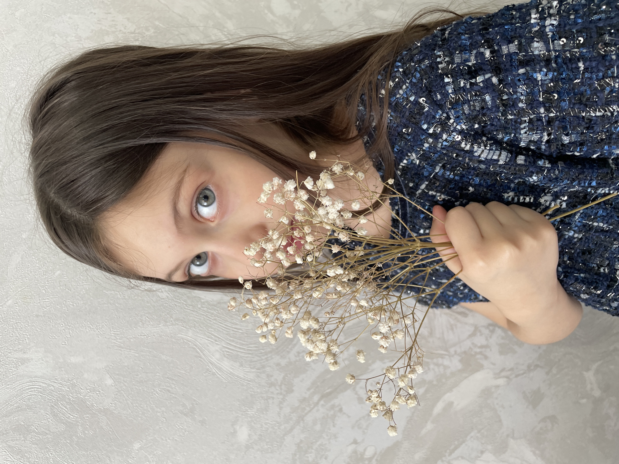 A young female, appearing to be a child, with long dark brown hair and light blue eyes, is depicted in a medium-close-up portrait. She is positioned against a light gray wall featuring a subtle, irregular textured pattern. The subject holds a small bundle of dried white flowers, resembling Gypsophila, with her left hand, positioning them to partially obscure the lower-right quadrant of her face, including her nose and mouth. She is wearing a dark blue, textured garment with interwoven white and silver threads or sequins. Her visible left eye is wide and directed towards the viewer. The specific location within Izhevsk, Russia, is not identifiable from the visual information provided.