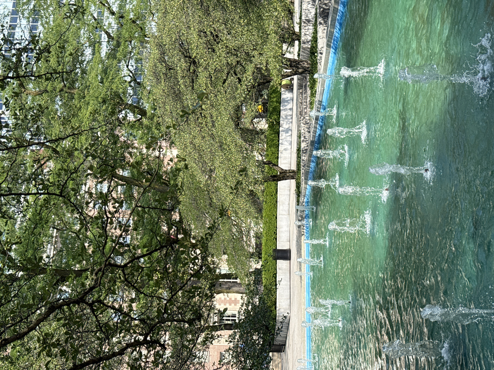 The image depicts a public fountain in a park-like setting with lush green trees and modern buildings in the background. The fountain is filled with turquoise water and features multiple jets of water spraying upwards, creating a dynamic and refreshing visual. The surrounding area includes paved walkways and manicured hedges, suggesting a well-maintained urban space. Sunlight filters through the leaves of the trees, casting dappled shadows on the ground. The architecture in the background suggests a contemporary urban environment. While the exact location isn't specified, the scene is characteristic of a public plaza or park found in a major city like Chicago, United States of America.
