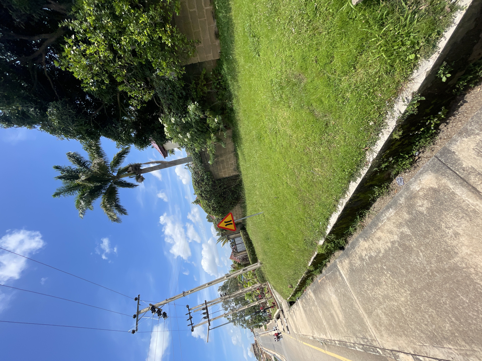 The image depicts a street scene in Jos, Nigeria, on a bright, sunny day. A concrete sidewalk runs along the left side of the frame, bordered by a yellow line on the road. Power lines and utility poles are prominently featured, stretching diagonally across the sky. Beyond the sidewalk, a well-maintained green lawn slopes upwards, with a brick wall and lush vegetation forming a backdrop. A traffic sign indicating a potential hazard, a split in the road, is visible on the lawn. In the distance, a few people can be seen walking along the road, and buildings are visible. A tall palm tree stands out against the blue sky dotted with white clouds, adding a tropical element to the urban landscape. The overall impression is one of a typical day in a Nigerian city.