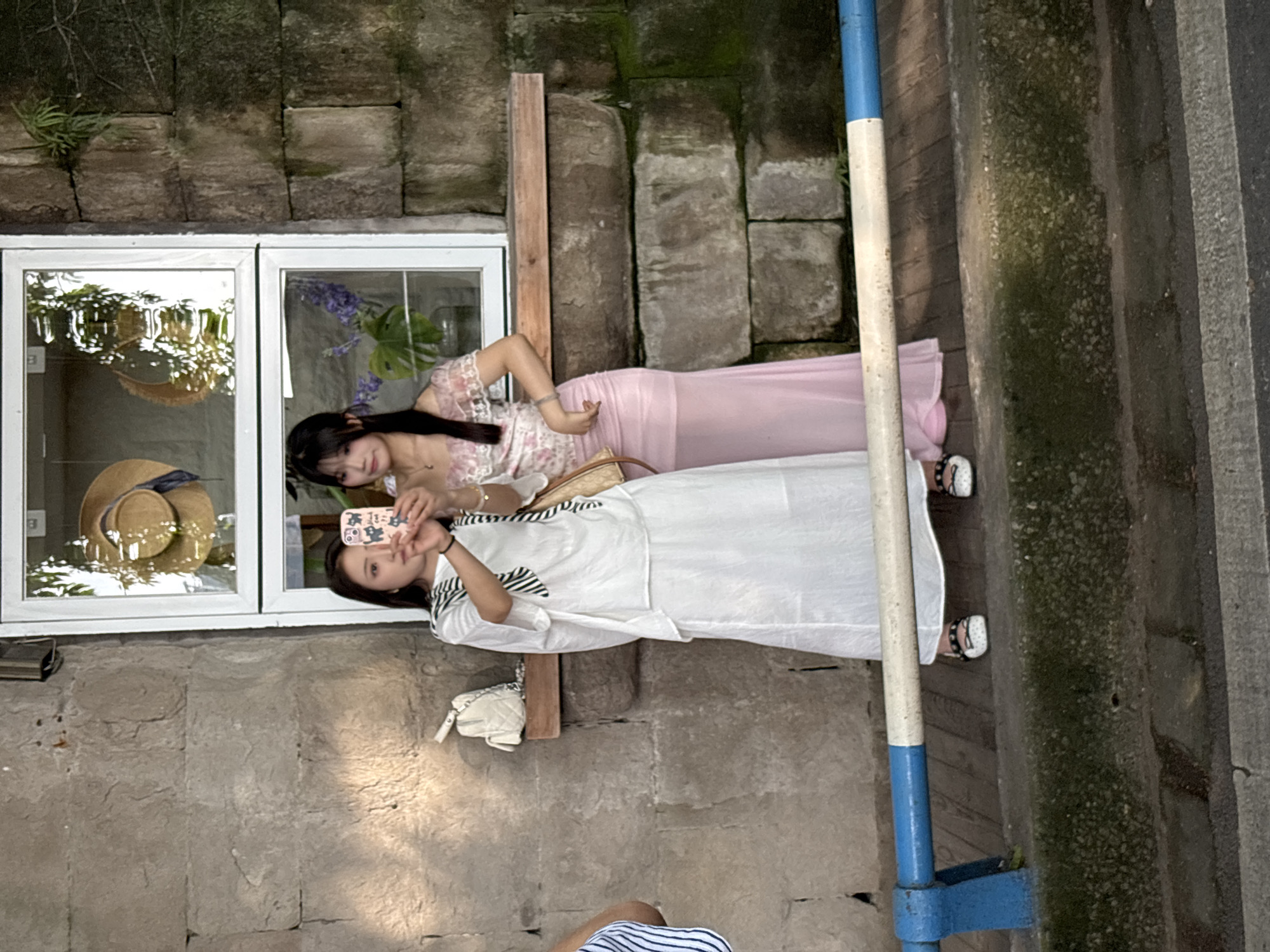 Two women pose in front of a storefront window in Yuzhong District, China. One woman in a white dress holds a phone and takes a photo of herself with the other woman who is wearing a pink dress. The storefront window contains a straw hat and floral arrangements. The building is made of stone and moss is present on some of the stones. A blue and white pole is on the right side of the frame.