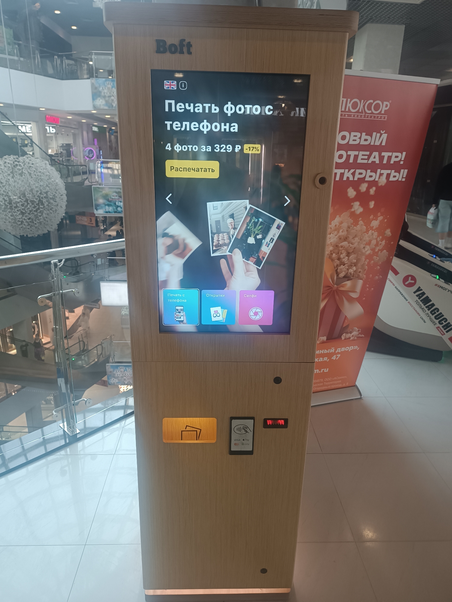 This is a full-shot, eye-level photograph of a Boft photo booth located inside a shopping mall. The photo booth is made of light-colored wood and has a touchscreen interface displaying options in Russian. The screen shows the Boft logo at the top, followed by text that translates to "Print photos from your phone." Below this, it indicates "4 photos for 329 ₽ -17%," suggesting a discount. The screen also displays options for "Print from phone," "Openings," and "Selfie," with corresponding icons. A yellow button labeled "Print" is visible below the offer. To the right of the photo booth, a large orange banner promoting "Luxury Cinema" can be seen. On the lower part of the photo booth, there is a card reader with "VISA" and "Pay" logos, as well as a small slot with illuminated red numbers. The background shows the interior of a shopping mall, including escalators and other retail spaces, with a bright, well-lit environment. The scene is devoid of any specific individuals interacting with the photo booth. The presence of the "Luxury Cinema" banner and the general mall environment suggests the location is likely a commercial area within Tula, Russia, although specific street addresses are not visible in the image.