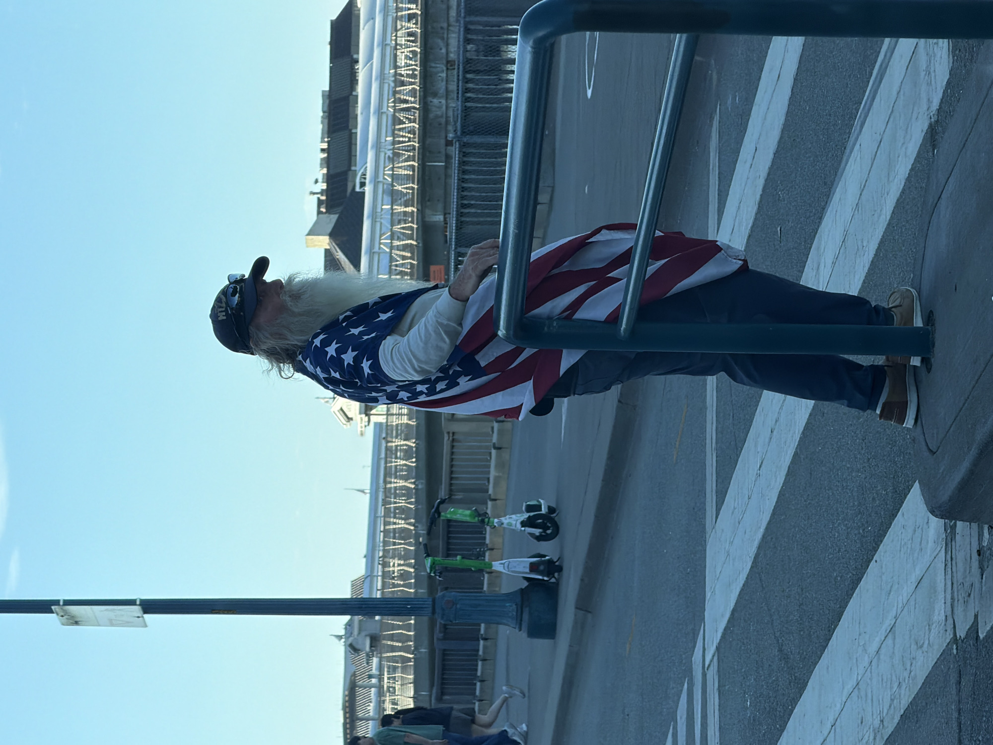 An older Caucasian male with a long white beard and a baseball cap is standing by a crosswalk. He is wearing a jacket or shirt adorned with an American flag pattern and dark pants. To his left, two electric scooters are parked next to a lamppost. In the background, there is a pedestrian bridge, buildings, and a clear blue sky. The scene appears to be an outdoor urban environment.
