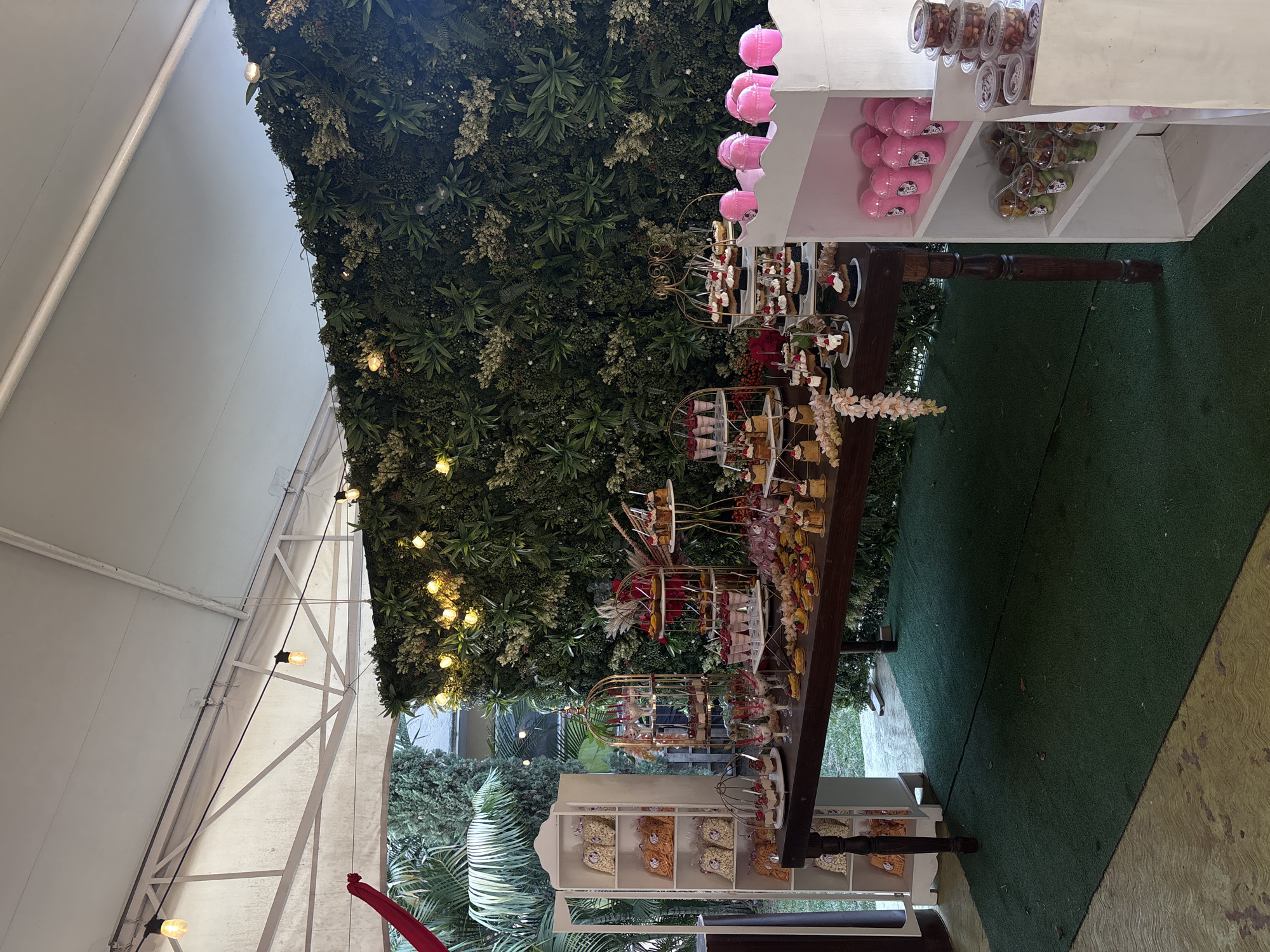 The image shows a dessert buffet under a white tent structure, possibly outdoors due to the surrounding greenery. The buffet is arranged on a dark wood table with white shelving units on either end. One unit displays pink stuffed animals, small clear containers of various items, and small plastic containers filled with green objects. The other unit displays bags of chips and other snacks arranged in individual cubbies. The table itself holds tiered stands filled with desserts, arranged in decorative cages. The backdrop is a wall of dense greenery, possibly artificial, adorned with string lights. In the background, a glimpse of trees and foliage can be seen. The floor is covered with green artificial turf. The location is potentially Chilpancingo, Mexico, although there is no visible textual or visual confirmation within the image.