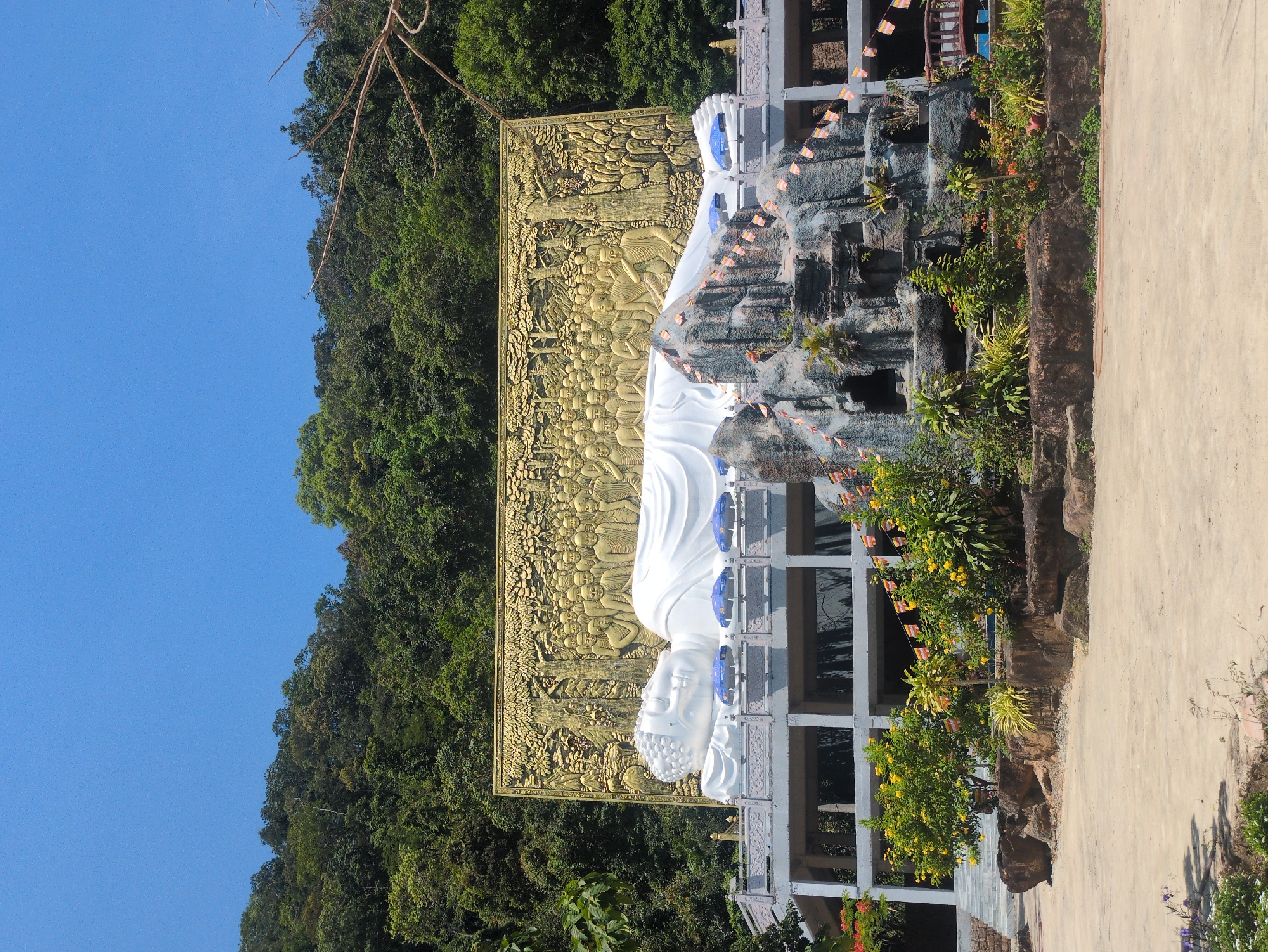 A horizontally oriented outdoor shot features a reclining Buddha statue situated in Phú Quốc, Vietnam. The statue is prominently white, juxtaposed against a large, golden rectangular backdrop intricately adorned with carved figures. The statue lies atop a multi-level structure of gray rocks and greenery, interspersed with architectural elements like columns and levels. Small flags are strung across the structure. Dense foliage flanks the statue on the left, transitioning into a clear blue sky. To the right of the statue is a pathway.