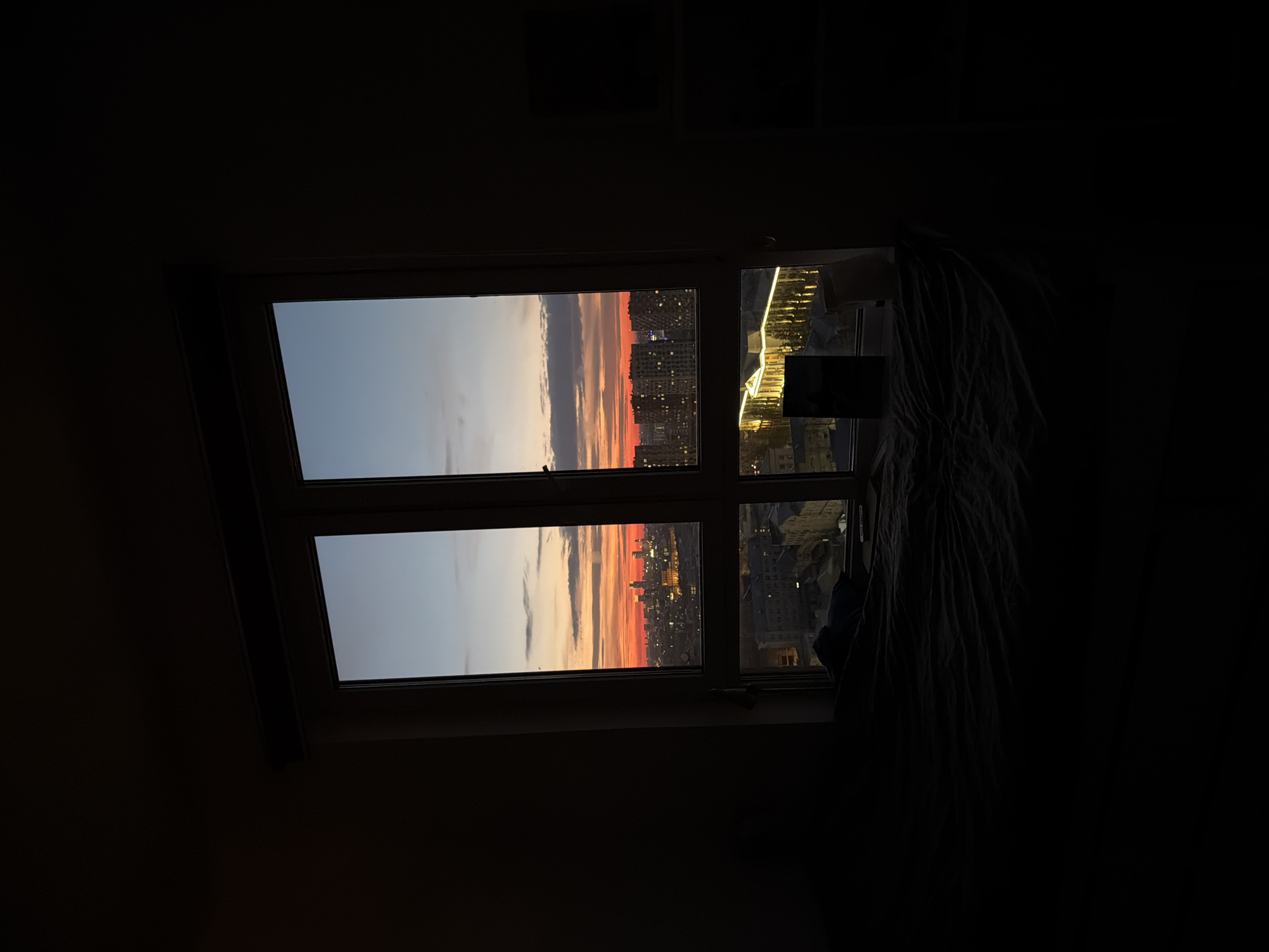 A window in a dark room frames a sunset over a city. The window is divided into multiple panes, two of which are open, revealing a cityscape silhouetted against a vibrant sky with orange and red hues. Other windows, situated on the right side of the image, show glimpses of brightly lit interiors and cityscapes. A curtain partially obscures the window on the right.