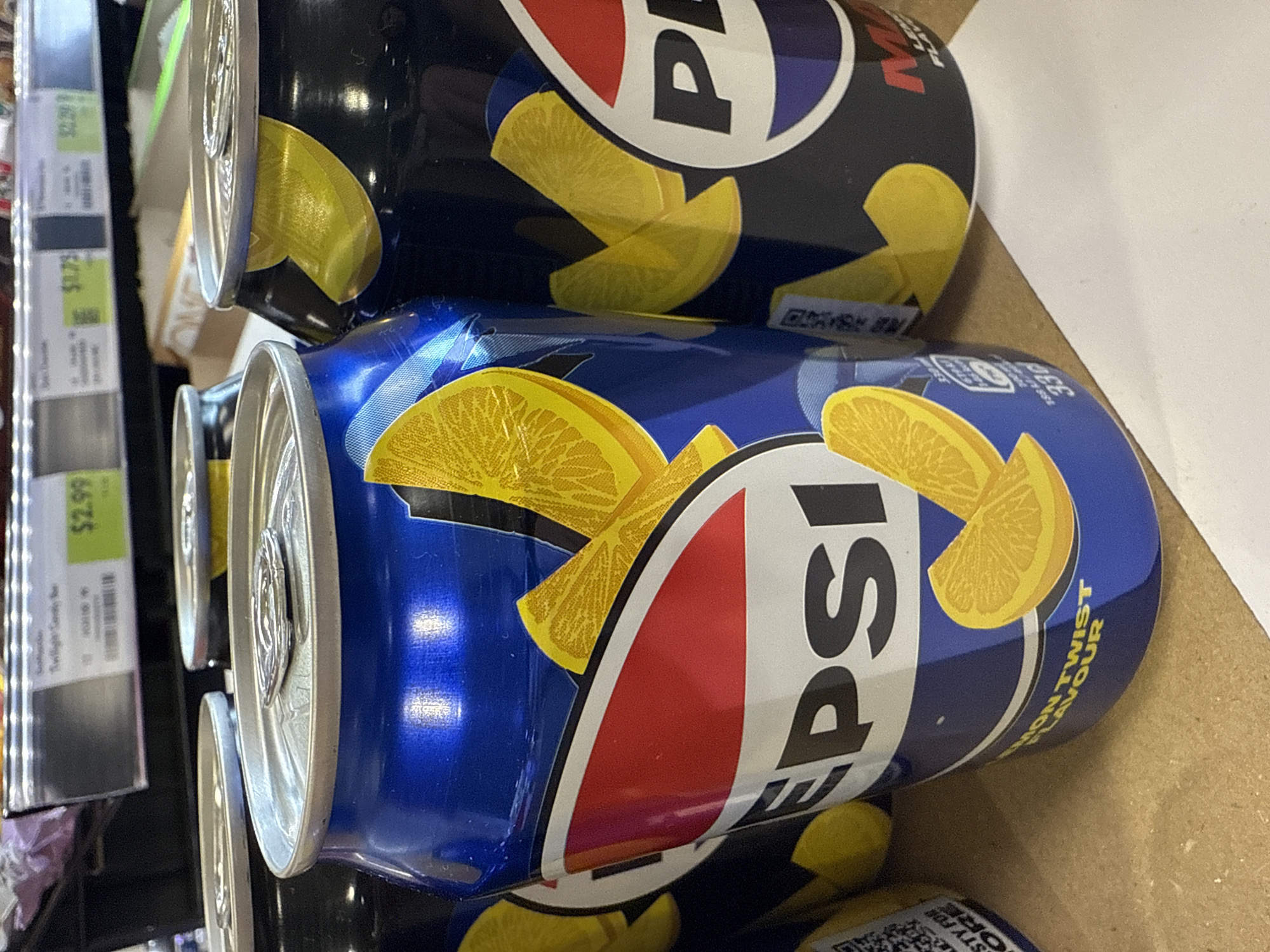 The image shows two cans of Pepsi Lemon Twist on a shelf, presumably in a store. The cans are blue and feature lemon illustrations. The shelf also contains a price tag reading "$2.99." The location is presumed to be in Darien, United States of America.