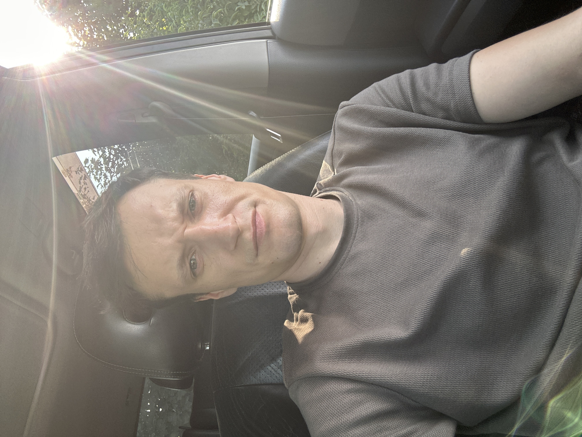 A man is pictured inside a car, seated in the driver's seat. He is looking towards the camera with a slightly furrowed brow and a neutral expression. The sun is shining brightly through the car's windows, creating lens flare and illuminating the interior. The man is wearing a grey t-shirt with a textured pattern. The car's interior is visible, including the leather headrest and seat. Outside the car, green foliage can be seen through the windows, suggesting a natural or suburban setting. The location of Shatava, Ukraine is not discernible from the image.