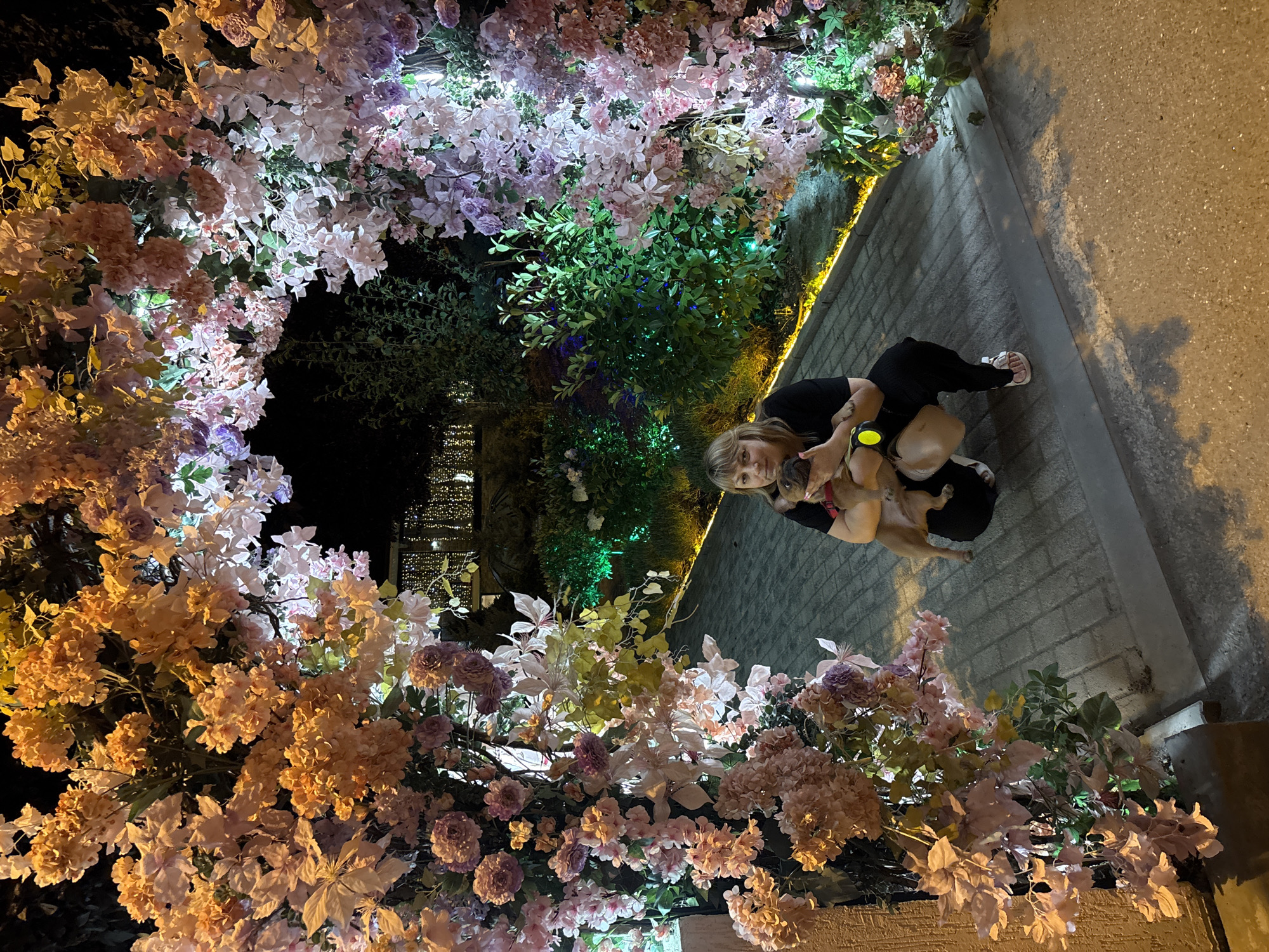 A woman kneels in front of a floral archway, holding a small dog. The arch is constructed from artificial flowers in shades of pink, peach, and purple, illuminated by artificial lights. The background shows a dark, outdoor environment with some greenery and string lights visible in the distance. The ground is paved with cobblestones. The woman is wearing a black outfit and white sandals. The overall scene appears to be an outdoor decorative display, possibly at an event or establishment. There is no visual information in the image to specifically place it in Krasnodar, Russia.