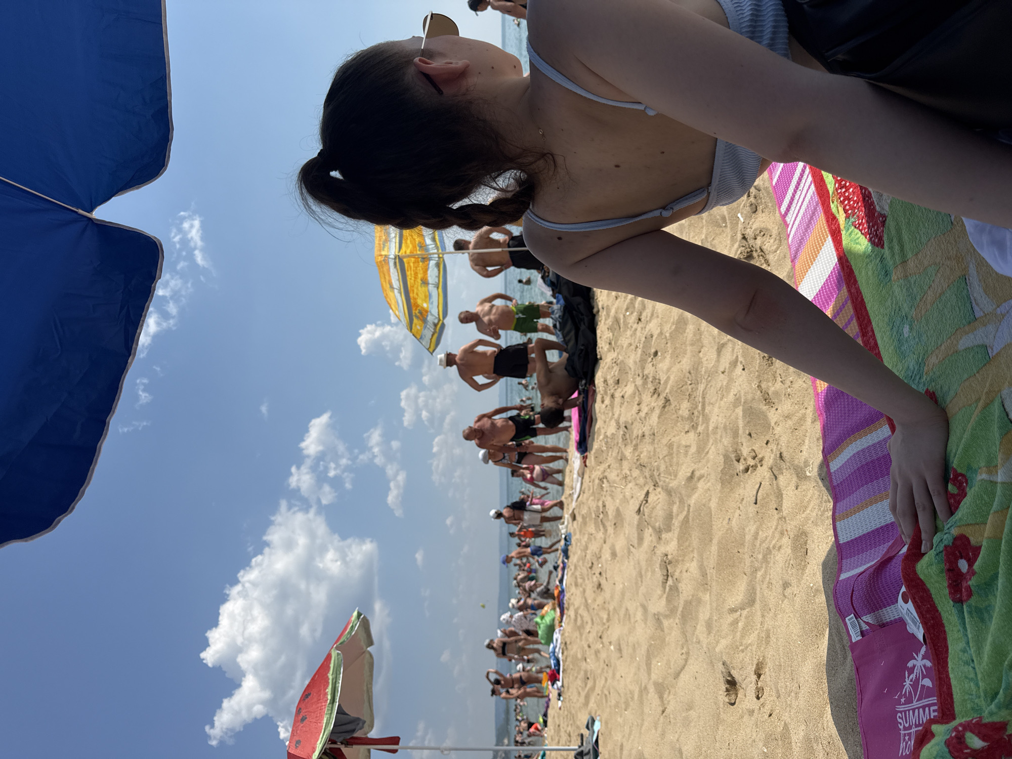 A crowded beach scene unfolds under a bright blue sky with scattered white clouds, likely at a Varna, Bulgaria beach like the Golden Sands or Sunny Beach, given the density of people and typical summer atmosphere. The foreground is dominated by a woman sitting on a colorful striped beach towel, her back to the camera, with her dark hair tied in a ponytail. She is wearing sunglasses and a light blue tank top. Her hand rests on the towel, which features patterns of flowers and palm trees. Behind her, the sandy beach teems with people, many in swimwear, some wading in the shallow water of the sea, others standing on the shore. Beach umbrellas, one shaped like a slice of watermelon and another yellow and white striped, dot the landscape, providing shade. Several men are visible in the middle ground, some appearing to converse. The sea stretches out in the background, meeting the horizon under the sunny sky.