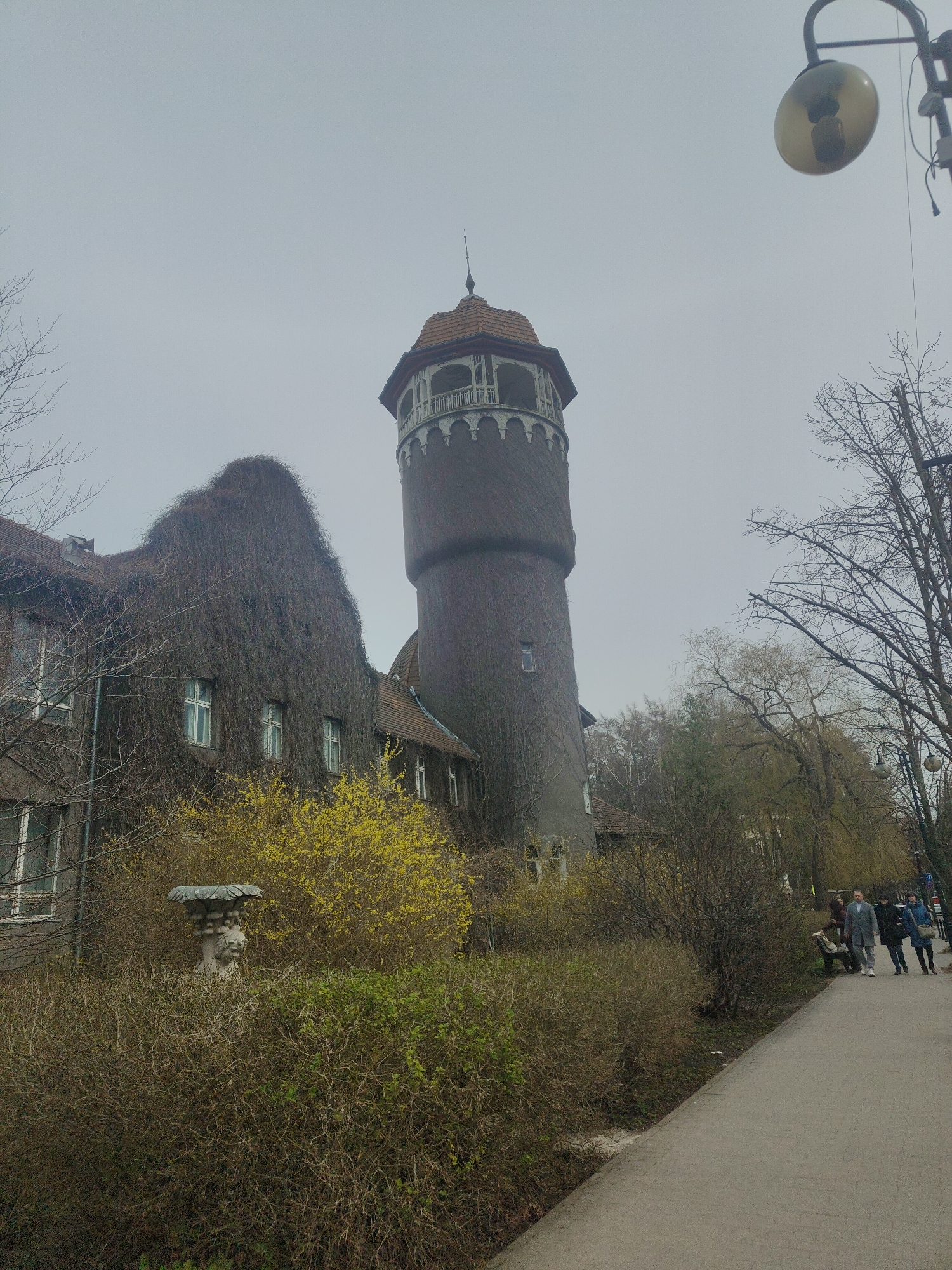 A medium shot of an overcast day depicts a water tower covered with ivy or similar climbing plant, next to a vine-covered building, in the city of Unknown, Unknown. The water tower is round with a red tile roof and a decorative balustrade near the top. The vine-covered building has several windows visible and appears to be a multi-story structure. In the foreground, there are overgrown bushes, some with yellow flowers, and what looks like a stone fountain or statue. A sidewalk runs along the right side of the image, and several people are walking along it. A lamppost with a round, yellow lamp is visible in the upper right corner.