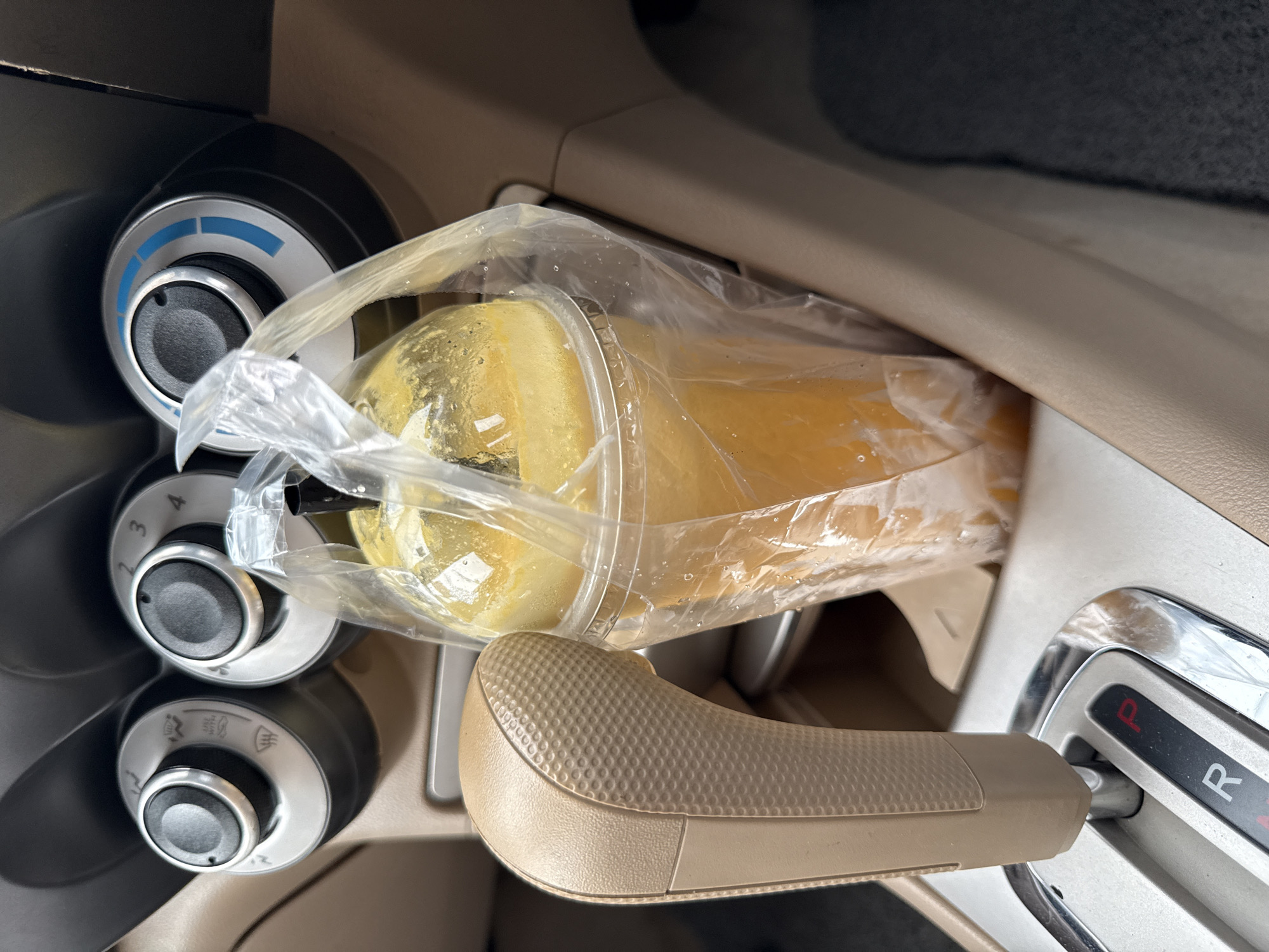 In Baan Bo Phut, Thailand, a transparent plastic cup filled with a vibrant orange beverage and ice is situated in a car's cup holder. The cup is enclosed in a larger, clear plastic bag, and a black straw extends from the top. The car's interior is visible, featuring a beige gear shift lever and part of the center console with climate control knobs. The scene appears to be captured during daylight.