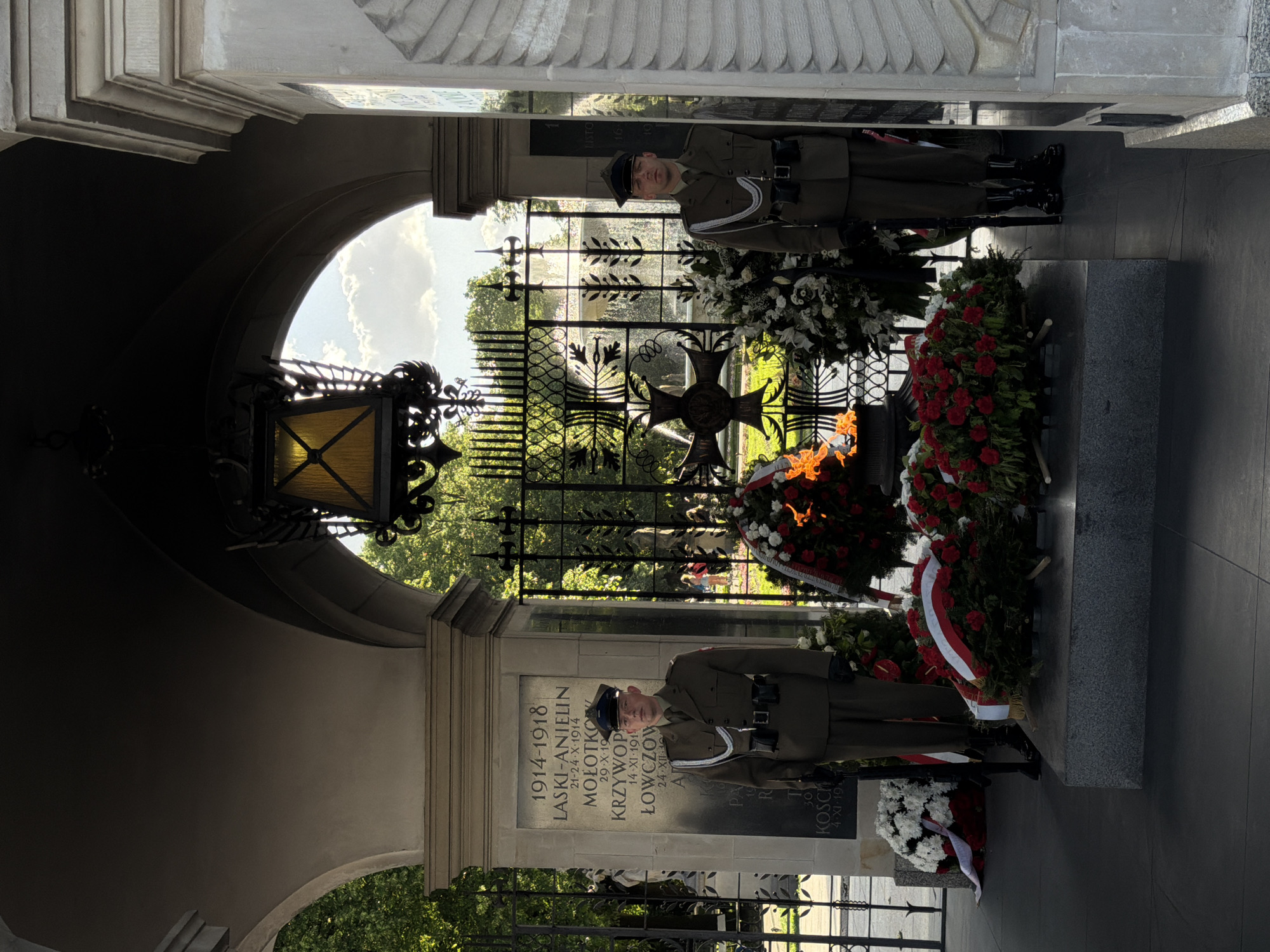 This image shows the Tomb of the Unknown Soldier in Warsaw, Poland, viewed through an arched structure. Two soldiers in uniform stand guard on either side of the tomb, which is adorned with wreaths and a flame. The tomb has an inscription, with "1914-1918" prominently displayed at the top. A decorative metal gate forms a backdrop, with trees and a building visible through the gate. The sky is partly visible through the top of the arch.