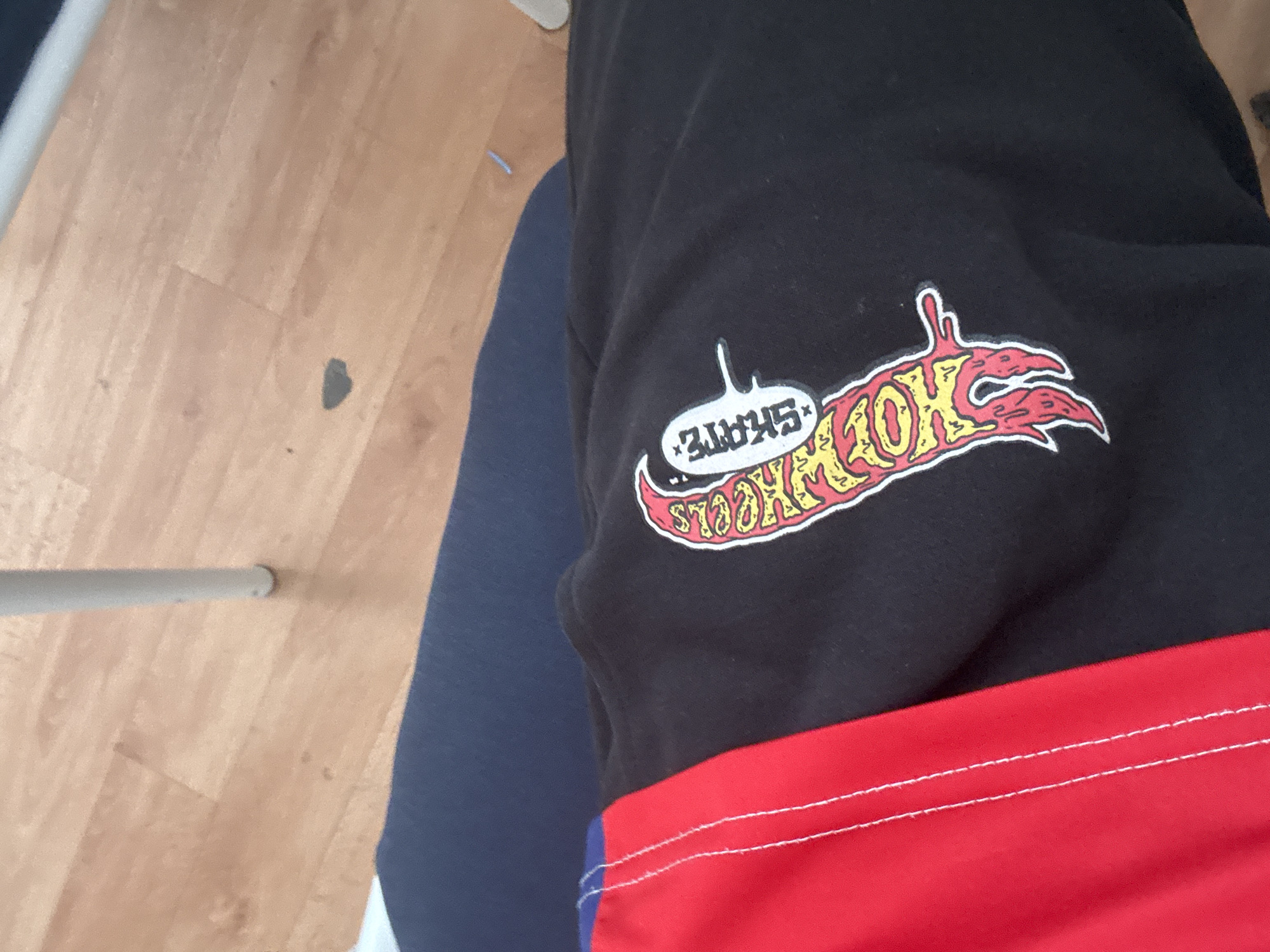 The image shows a close-up of a black garment with a red and blue striped panel on the left side. A graphic design featuring the word "SKATE" in a bubble shape above a flame-like illustration is present on the black portion of the garment. The background appears to be a wooden floor with a white chair leg visible. The scene is indoors.