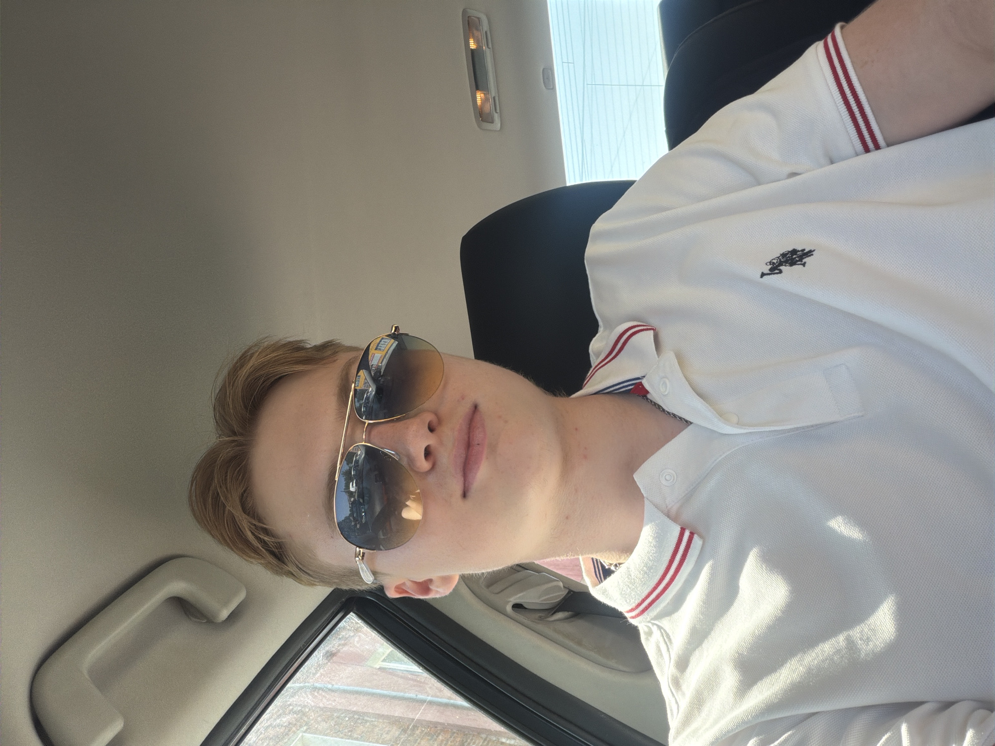 A young man with blond hair and sunglasses is seated inside a vehicle, looking towards the camera. He is wearing a white polo shirt with red and blue striped trim on the collar and sleeves, and a small embroidered logo on the left chest. The interior of the car is visible, including a handle on the ceiling and the top of a headrest. The scene appears to be a sunny day, with light reflecting on the man's shirt and the car's interior. The background outside the car window shows a brick building and trees, suggesting an urban or suburban setting. Based on the provided information, this image was captured in Polonne, Ukraine.