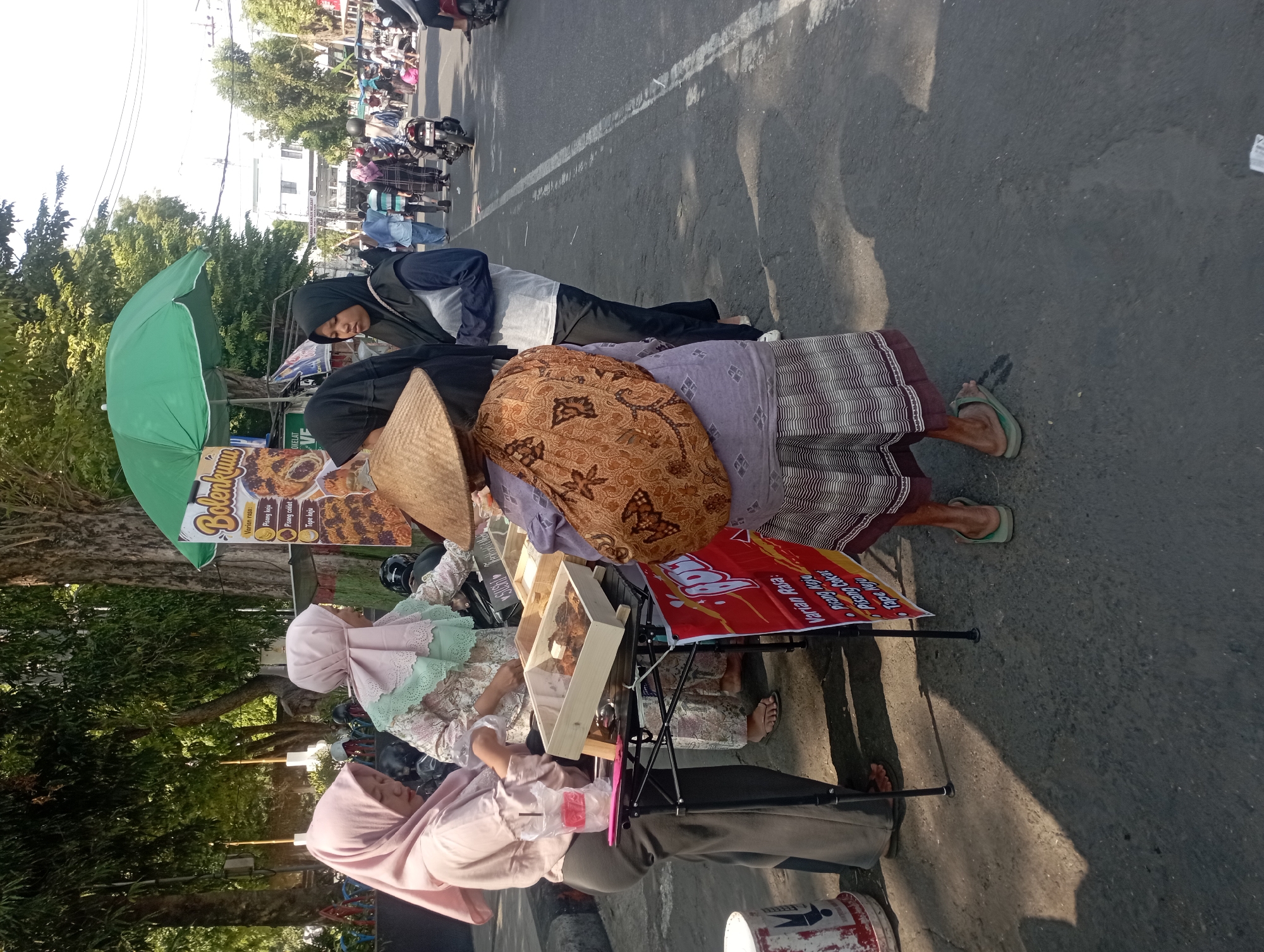In Nganjuk, Indonesia, on a sunny day, a street vendor is selling baked goods from a stall set up on the side of a road. The vendor, wearing a conical hat and a patterned bag slung over her shoulder, stands behind a display of food items. Several other women, some wearing hijabs, are gathered around the stall, interacting with the vendor. A green umbrella provides shade over the stall. In the background, other street vendors, pedestrians, and a motorcycle are visible. The scene suggests a bustling marketplace or a busy street in a city.