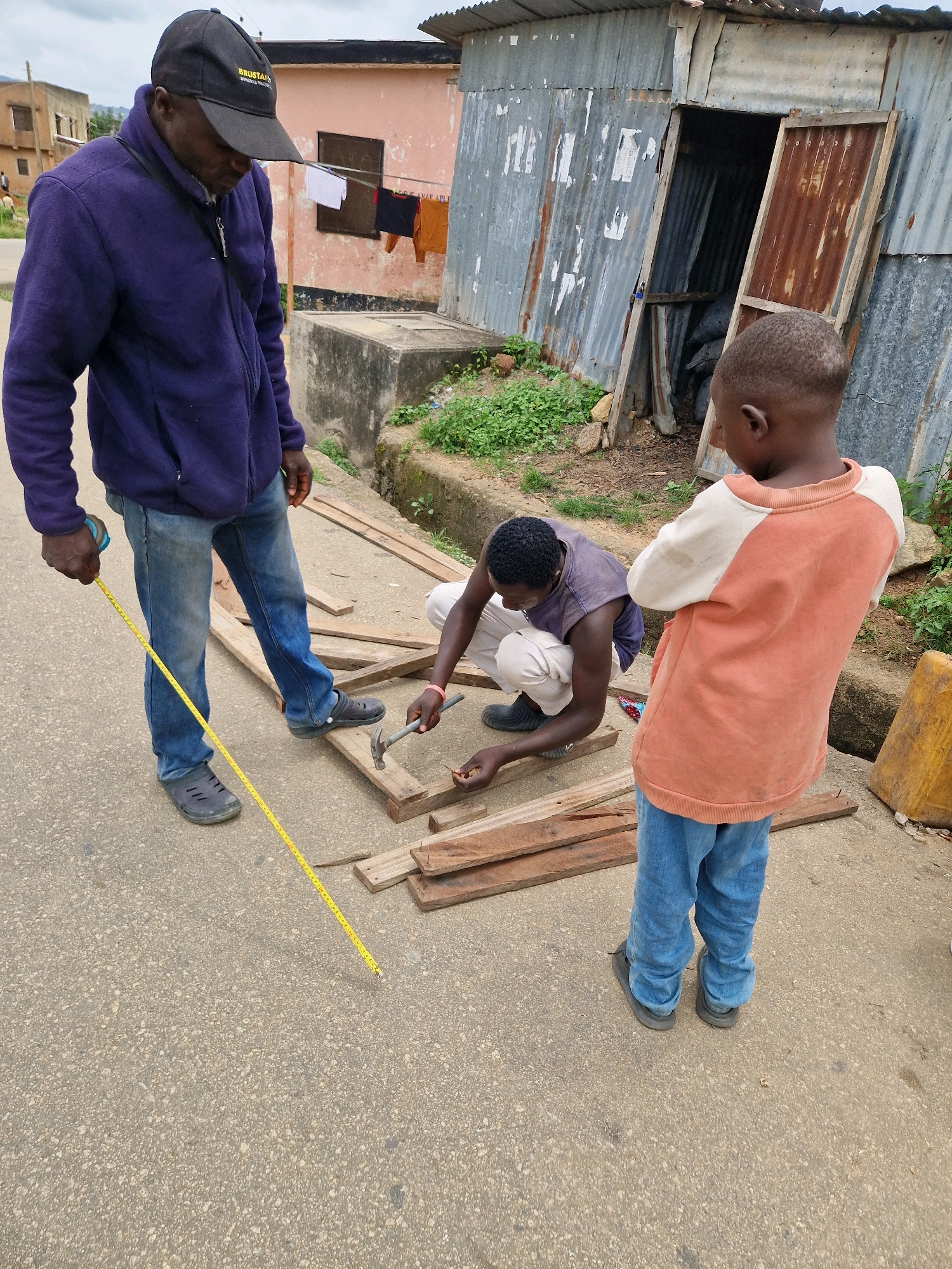 In an outdoor urban setting, three individuals are engaged in what appears to be construction or repair work. A man wearing a blue jacket and a black baseball cap is holding a yellow measuring tape, extending it across a paved surface. Next to him, another man in a purple tank top and light-colored pants is kneeling and using a hammer on wooden planks. A young boy in an orange and white long-sleeved shirt and blue jeans stands observing the activity. The background features a corrugated metal structure with a wooden door, a pink building, and some greenery. The overall scene suggests a practical, hands-on task being carried out in a modest urban environment.