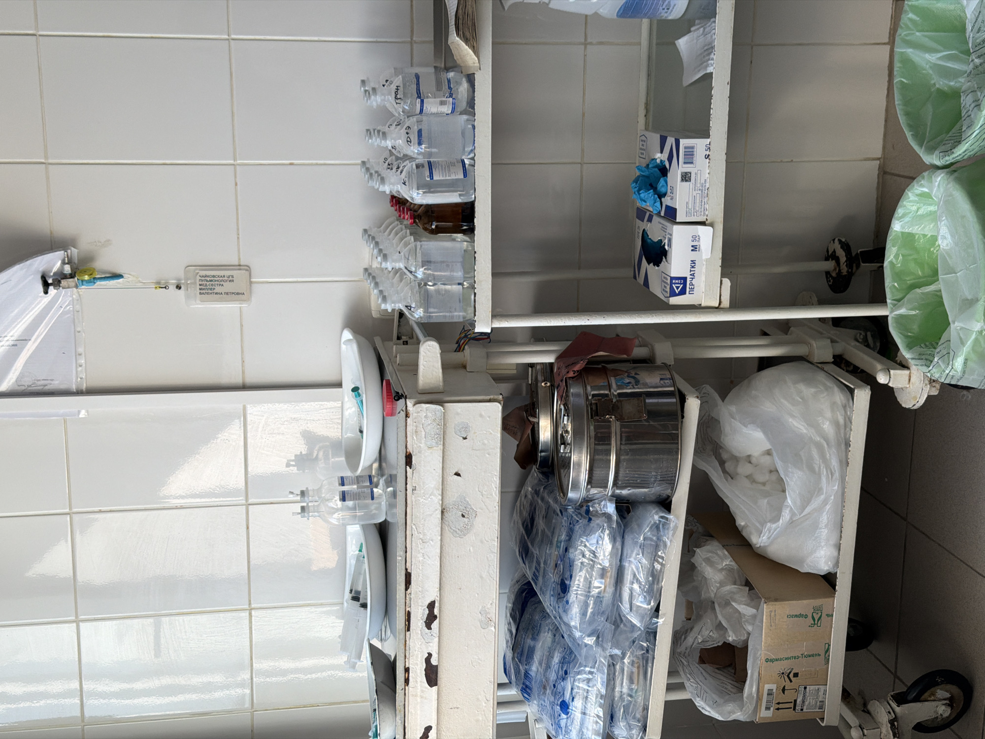 The image shows a storage area in what appears to be a medical facility, indicated by the text “Chaikovskaya Central City Hospital, Pulmonology". It is located in the city of Chaikovsky, Russia. The shelves contain various medical supplies, including IV fluid bags, boxes of gloves, sterilization containers, bags of cotton, and other wrapped medical items. The presence of oxygen tubing and other medical apparatus suggests a hospital or clinic setting. The tile wall background indicates a clean, sterile environment.