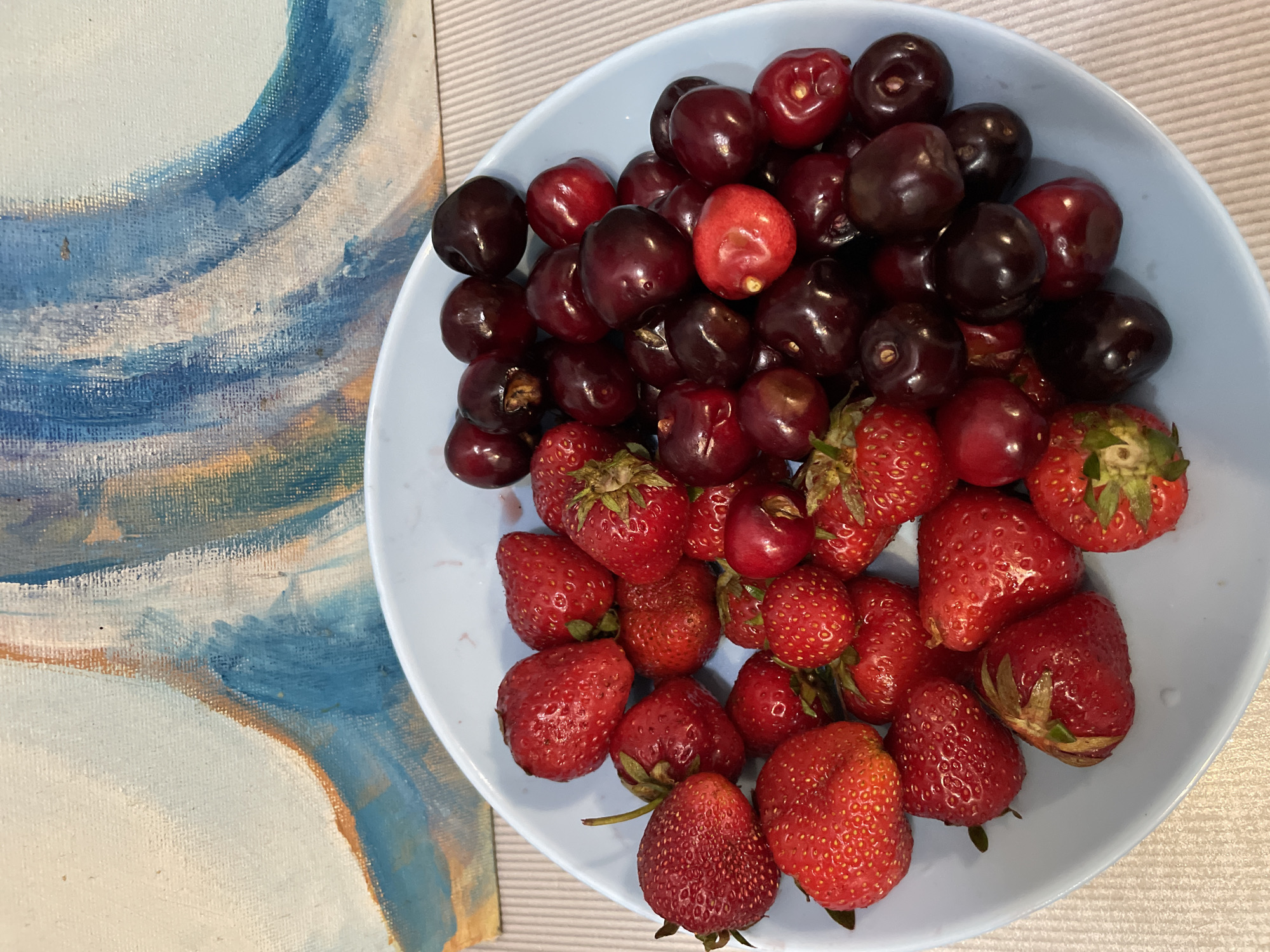 A light blue ceramic bowl filled with fresh strawberries and cherries sits on a light gray textured surface. The bowl is positioned in front of a canvas painting that features abstract brushstrokes in shades of blue, white, and orange. The painting has a textured, canvas-like appearance. The lighting suggests an indoor setting. The scene is devoid of any individuals or specific landmarks to indicate a precise location within the city of Sochi, Russia.