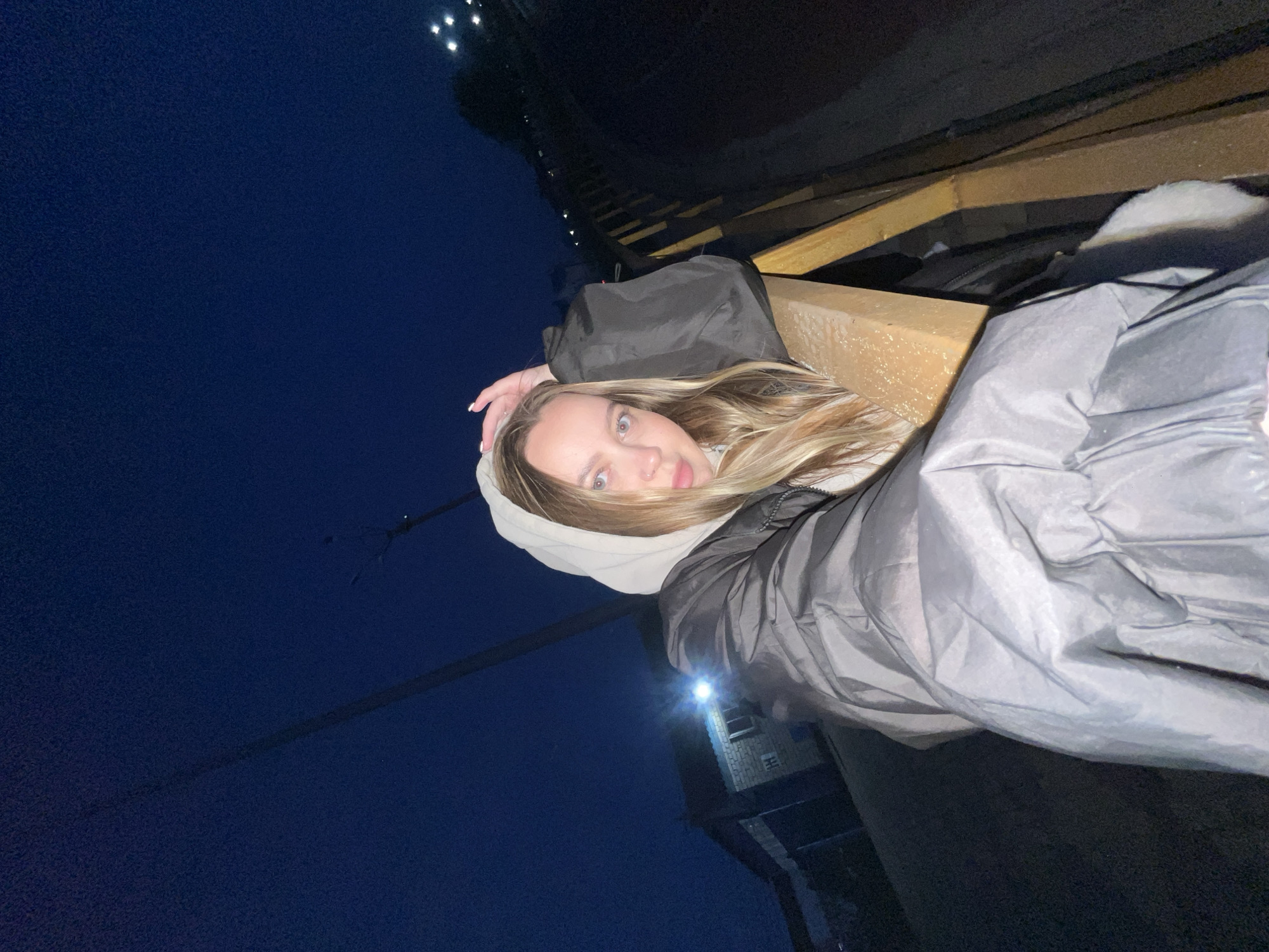 The image is a rotated portrait of a young woman taken at night, creating a unique perspective where she appears to be lying on her side or leaning back on an angled structure. She has long, wavy blonde hair, blue eyes, and fair skin, looking directly into the camera with a neutral expression. She is dressed in a light-colored hooded sweatshirt beneath a dark grey or black puffer jacket, suggesting cool temperatures. Her hand, adorned with light-colored nail polish, rests near her head.
The setting appears to be an outdoor or partially enclosed elevated space. She is positioned against and partially within a structure made of wooden beams or planks. The background consists of a dark blue, night sky, indicating the time of day. Several bright white lights, possibly streetlights or distant building lights, are visible against the dark sky in the upper left. A large, dark building, possibly industrial or an older structure, is faintly discernible in the lower right background, with one bright light illuminating a small section of it. The location context is Novopokrovskaya, Russia. There is no visible text in the image. The primary activity is the woman posing for the photograph.