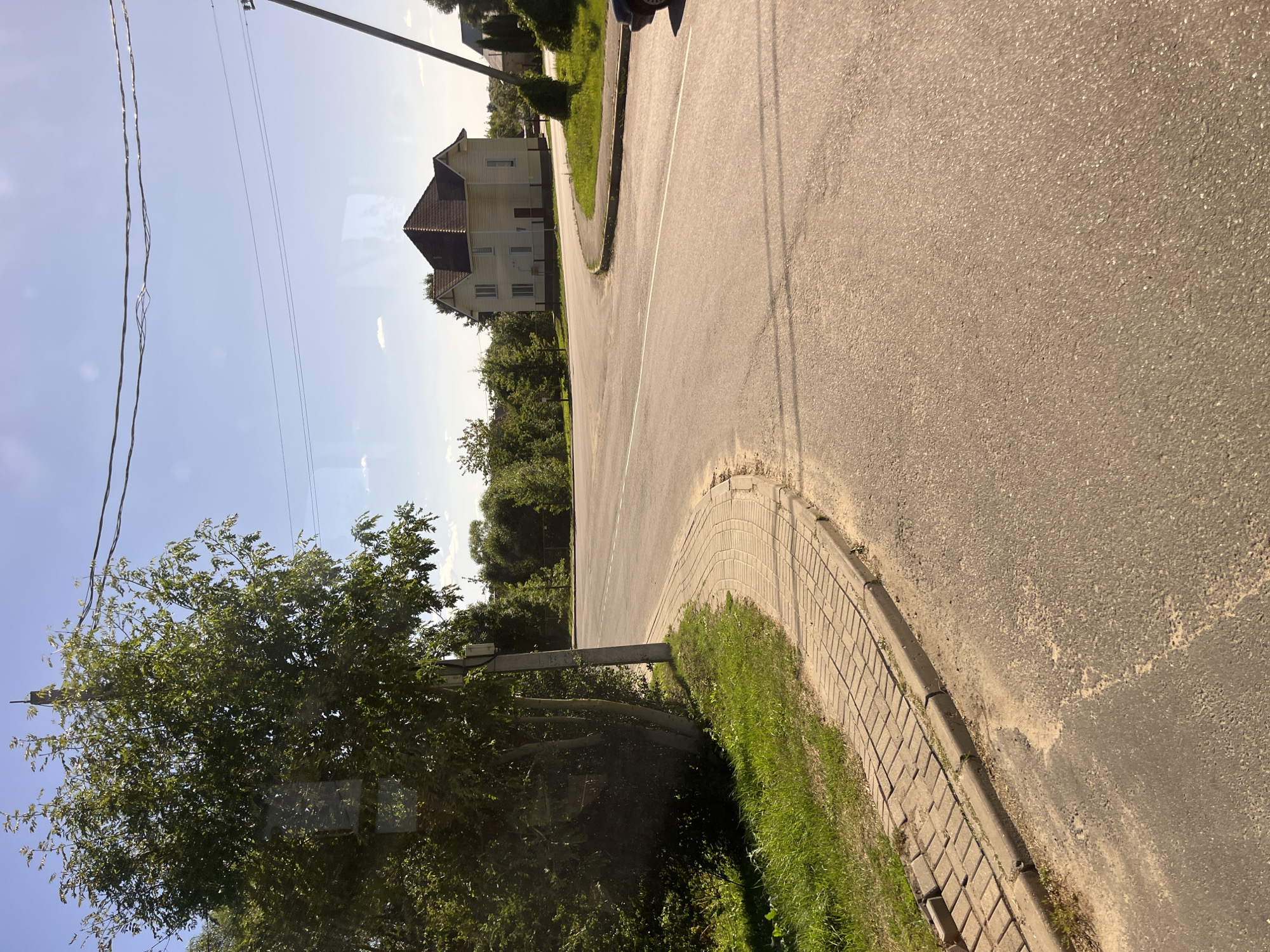 The image captures a daytime scene at a road intersection, likely in Pastavy, Belarus, given the architecture and environment. A paved road curves through the frame, with a section of brick sidewalk visible along the left edge. A mature tree with dense green foliage stands to the left, partially obscuring a utility pole. In the background, a light-colored, two-story house with a dark tiled roof is situated on a grassy area. Additional trees and foliage are visible behind the house, and power lines crisscross the bright blue sky. A dark-colored car is partially visible on the right side of the road. The sun casts shadows, indicating a bright, sunny day. The scene appears to be in a semi-rural or suburban area.