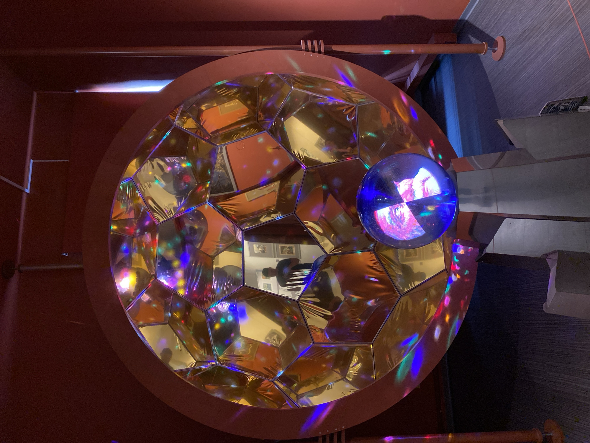 This image depicts a multifaceted, spherical object with a golden, reflective surface, reminiscent of a geodesic dome or a disco ball. The surface is composed of numerous geometric panels, each reflecting colored lights and distorted images of the surrounding environment. In the center of the reflection, a person, presumably the photographer, is visible wearing a dark t-shirt and holding a phone. Several framed pictures are also visible on the wall behind the reflected person.
In front of the large golden sphere, a smaller, transparent blue sphere rests on a metallic pedestal. This sphere also displays a fragmented image, appearing to be a close-up of a person's face. The background is a deep red color, with what appear to be industrial-style light fixtures or structural elements on the right side of the frame. The floor is covered with a patterned carpet in shades of gray and purple.
While the image shows an indoor installation, it does not provide any specific visual cues that would allow for identification of its location in Petrozavodsk, Russia. The context of the artwork or exhibition is also unknown.