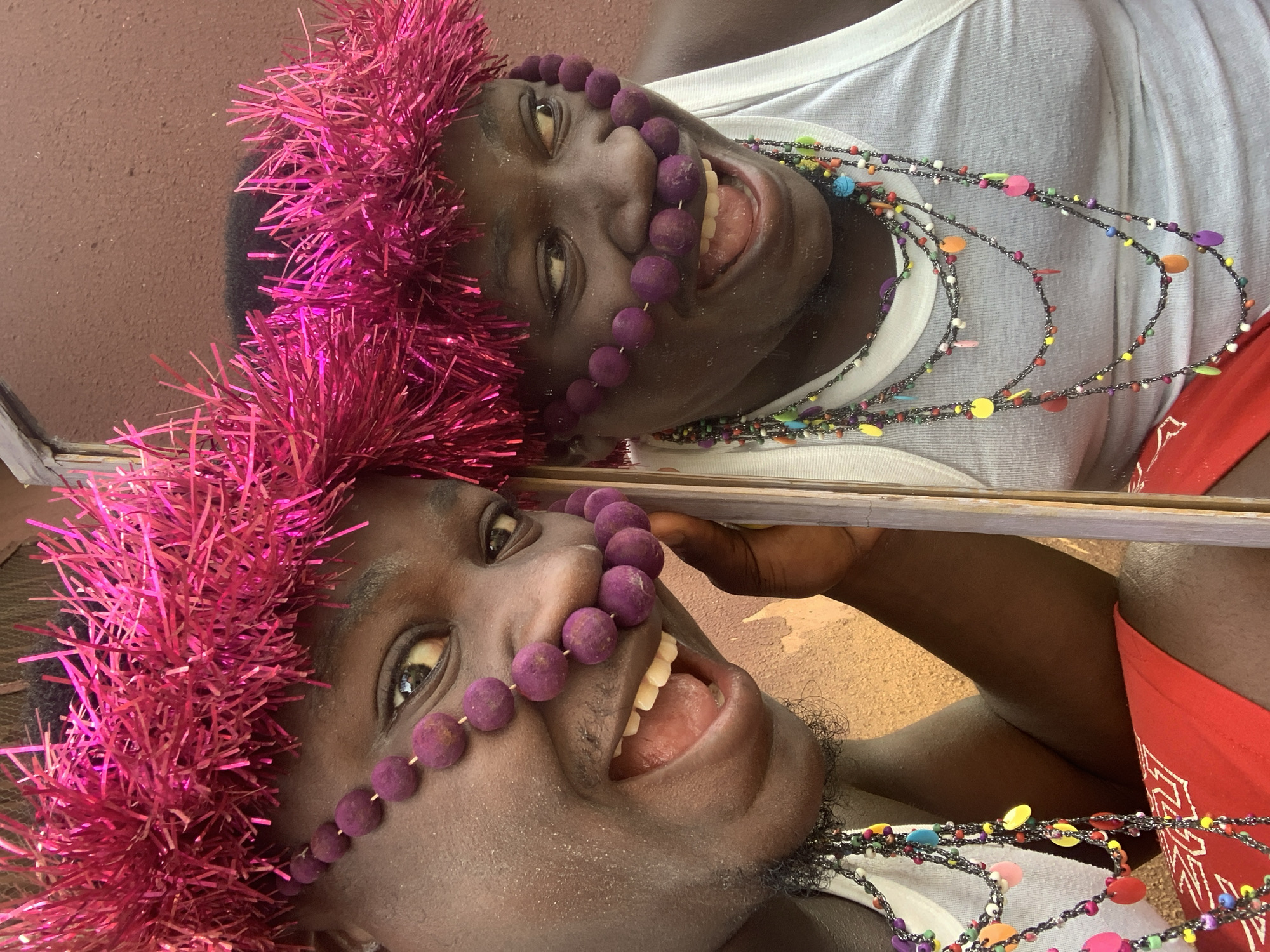 A close-up, waist-up portrait of a young Black individual is captured in Akwanga, Nigeria. The individual is adorned with a vibrant pink tinsel headdress and multiple strands of colorful beaded necklaces, some of which are woven through their nose. They are wearing a white tank top and a red garment, possibly a bandana, visible at the bottom. The individual is smiling broadly, with their mouth open and tongue visible. They are looking towards the right side of the frame. The background is a textured brown wall. The image is a selfie, as indicated by the reflection of the individual in what appears to be a mirror or reflective surface. The lighting suggests an outdoor environment during the day.