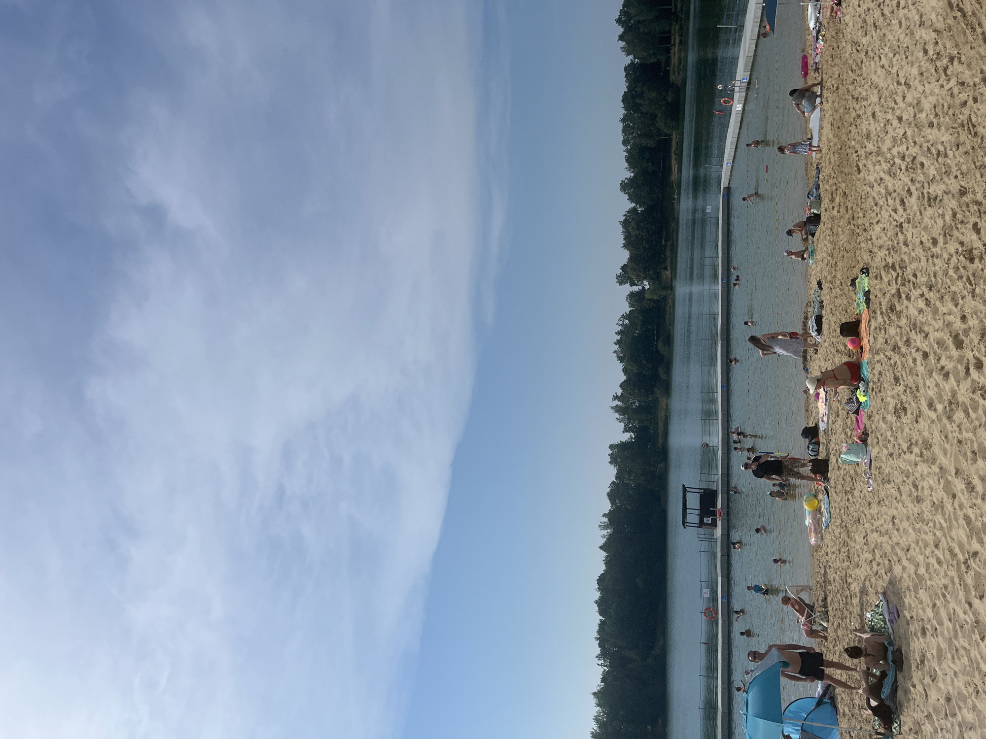 This is a wide shot of a crowded beach next to a swimming area. The scene is divided horizontally, with the left side dominated by a bright blue sky with wispy clouds, and the right side showing a sandy beach with many people and a large body of water.
The beach is the primary focus in the foreground, with numerous individuals scattered across the sand, some lying down on towels, others sitting or standing. Beach accessories like towels, bags, and a blue umbrella are visible. The water appears to be a swimming pool or a calm lake, with a white border and some dividing ropes. Several people are in the water, swimming or wading. There are also trees lining the far edge of the water, suggesting a natural setting.
The activity is clearly recreational, with people enjoying a sunny day at the beach and in the water. The lighting suggests it is daytime, likely midday or afternoon, given the bright sky and shadows. There are no discernible signs of text in the image. The overall mood appears relaxed and festive, typical of a summer day at a public swimming area.