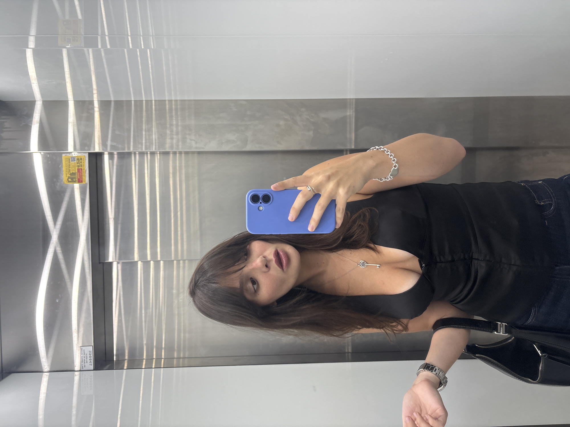 A young woman with long brown hair and bangs, wearing a black V-neck top and blue jeans, takes a selfie in an elevator. She is holding a blue smartphone in her right hand, capturing her reflection. She wears a silver necklace with a key pendant, a silver ring on her right index finger, and a silver bracelet on her left wrist. A black handbag hangs from her left arm. The elevator's interior is lined with reflective stainless steel panels and features white vertical accents. A yellow warning sticker is visible on the left panel. The image is a candid shot, likely taken in a residential or commercial building in Tel-Aviv, Israel.