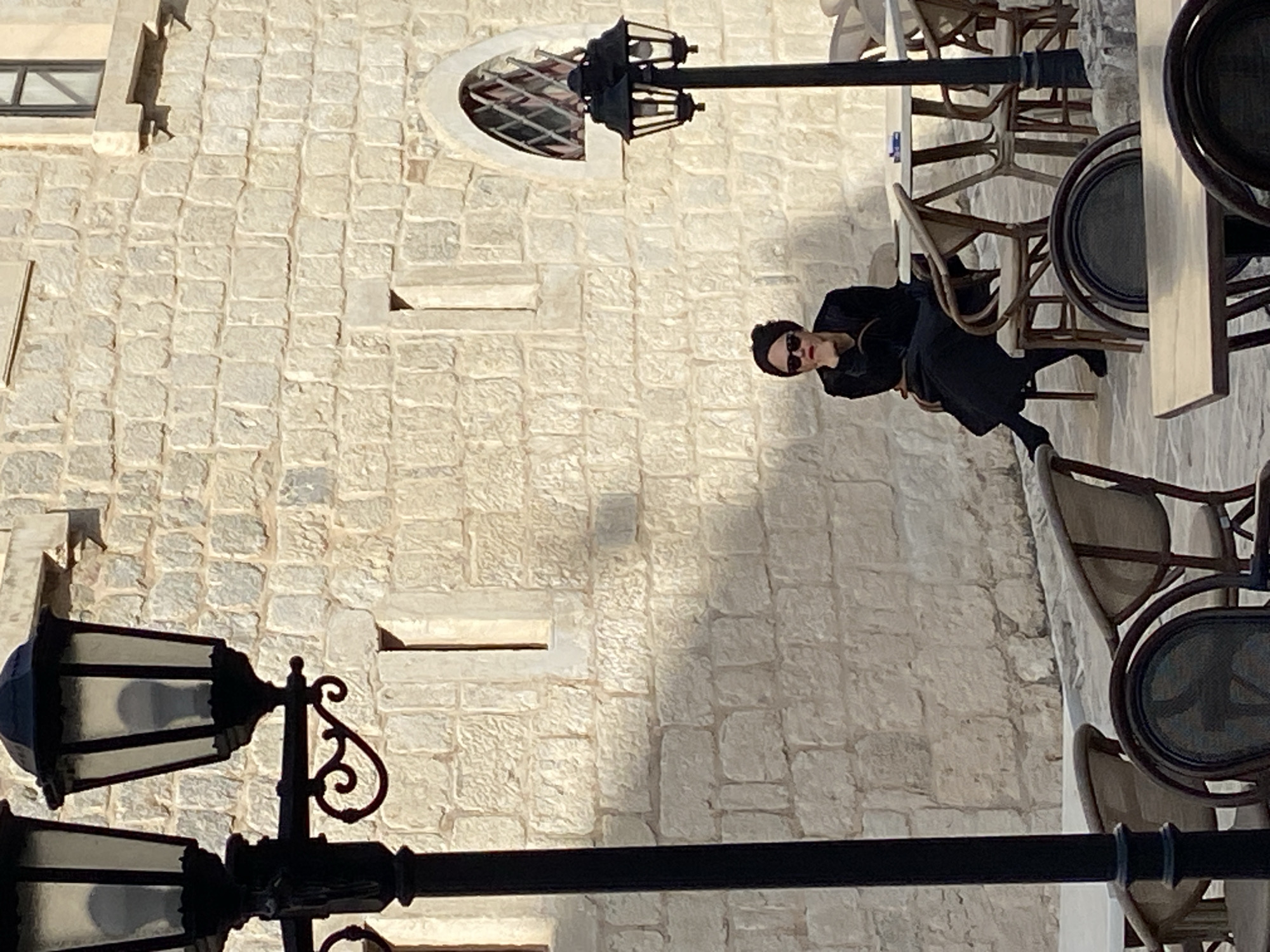 A woman in black clothing and sunglasses sits at an outdoor cafe in Budva, Montenegro. The cafe has light-colored stone walls and black iron lampposts. There are numerous chairs and tables visible. The cafe's seating arrangement suggests it is situated along a narrow street or alleyway.