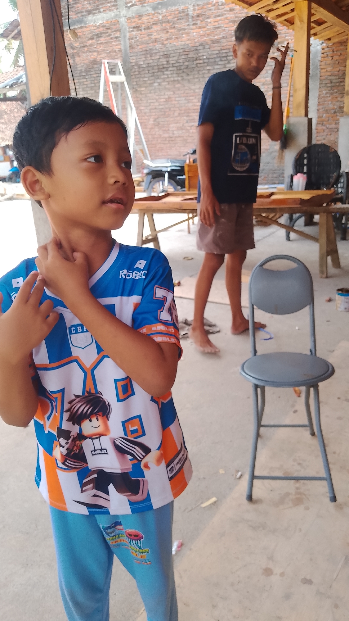 In Karanglangu, Indonesia, a young boy wearing a blue and white t-shirt featuring a cartoon character and blue pants stands in the foreground. His right hand is raised to his neck. Behind him, a teenage boy in a dark blue t-shirt and brown shorts stands with his right hand near his face. A gray folding chair is to the right of the teenage boy. The scene appears to be outdoors, with a brick wall in the background and wooden tables and structures suggesting a workshop or construction area. The ground is concrete.