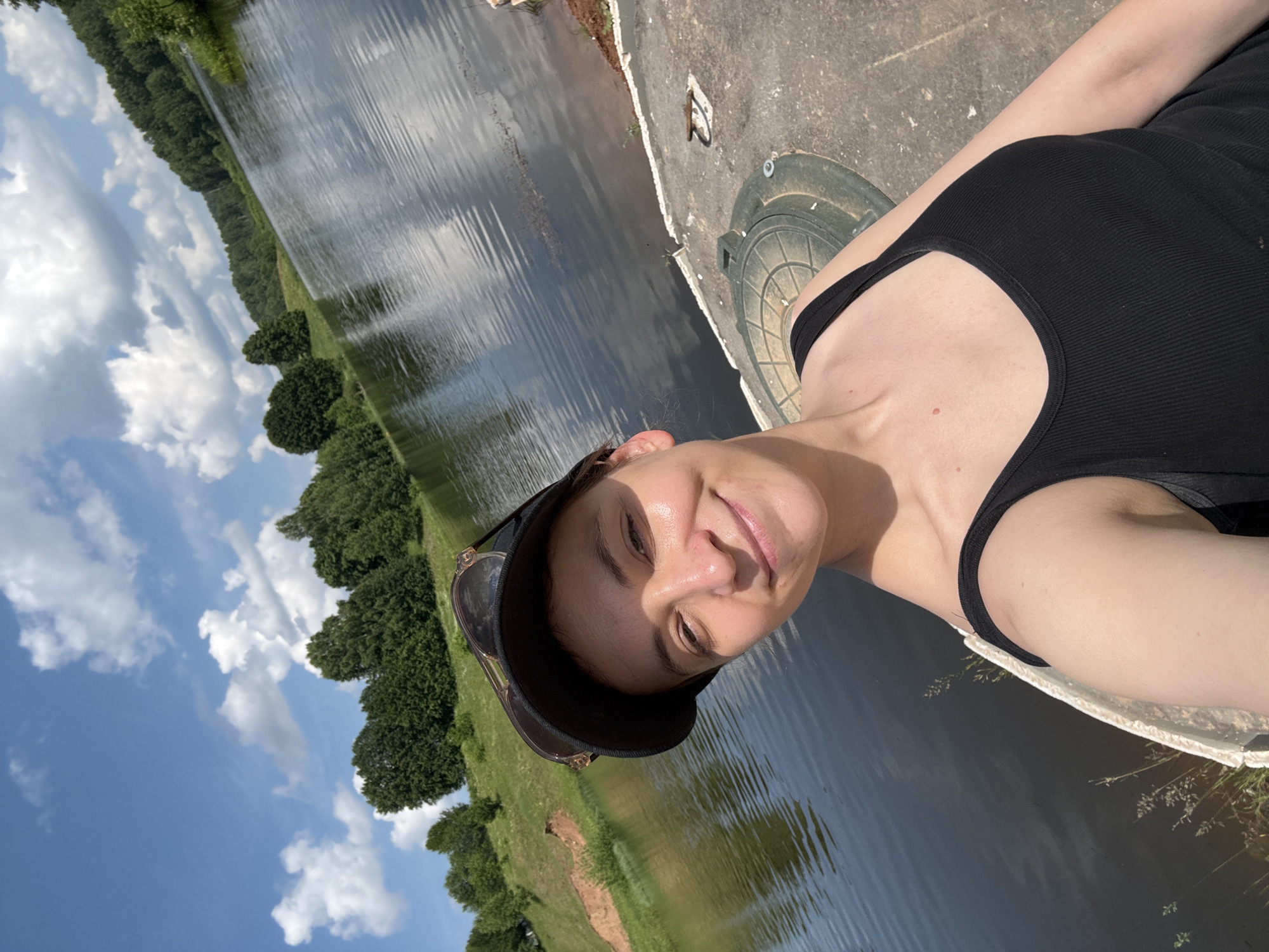A young woman wearing a black tank top and a black baseball cap with sunglasses perched on top is taking a selfie by a body of water. She has a bright smile and her face is illuminated by the sun. In the background, lush green trees line the shore of a calm river or lake. The sky above is a vibrant blue with scattered white clouds. A concrete platform is visible in the foreground, with a green circular hatch cover. The scene appears to be a serene outdoor setting in Zasechnyi, Russia.