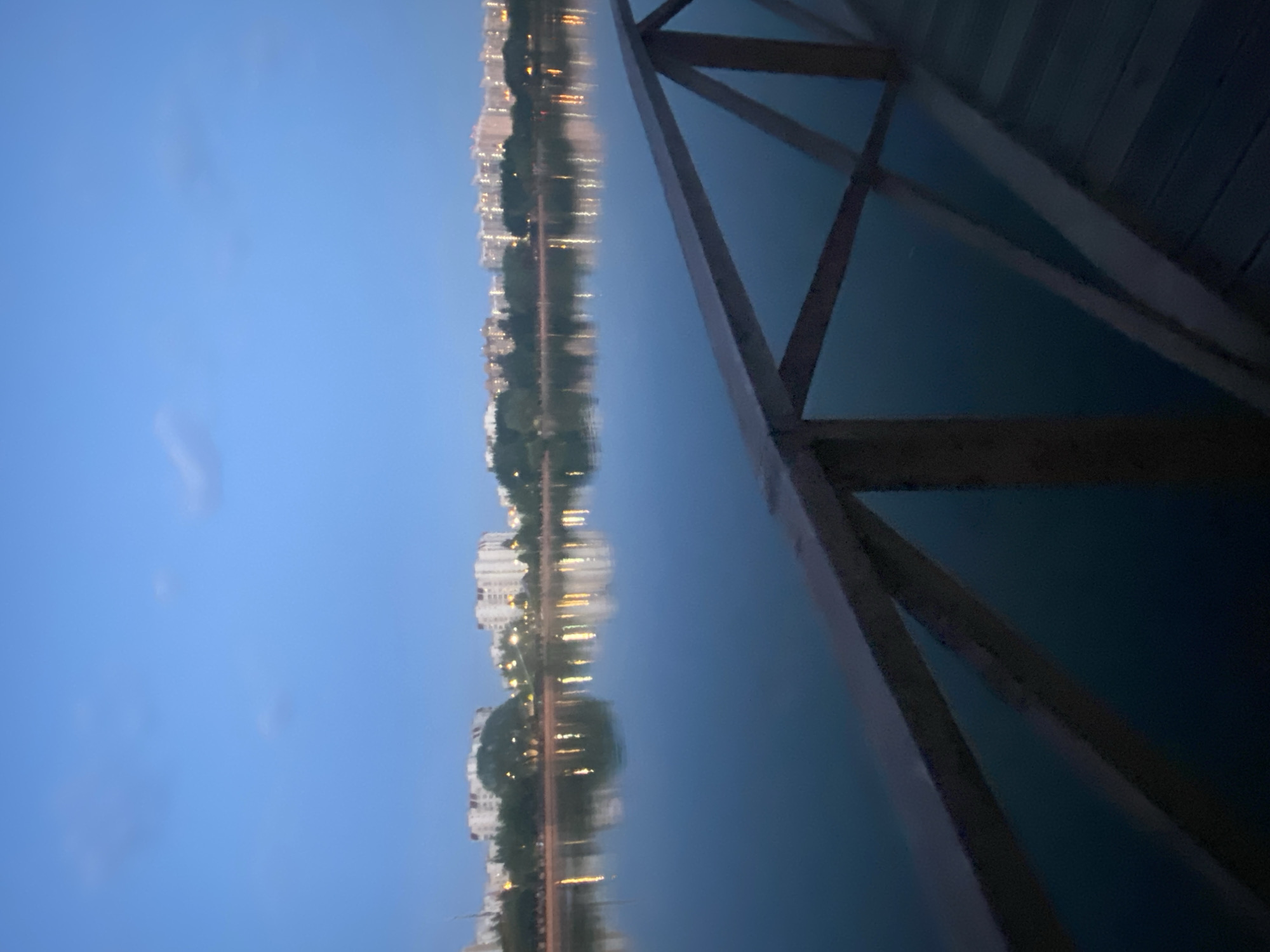 The image shows a body of water reflecting a cityscape at twilight. The water is dark and still, with a clear reflection of buildings, trees, and lights. On the right side of the image is a wooden structure, possibly a pier or dock. The sky is a gradient of blue, indicating the time of day is late afternoon or early evening. The image suggests a location in a city with a lake or riverfront area, possibly in Moscow, Russia, given the architectural style of the buildings in the reflection. The scene is quiet and serene, with no people visible.