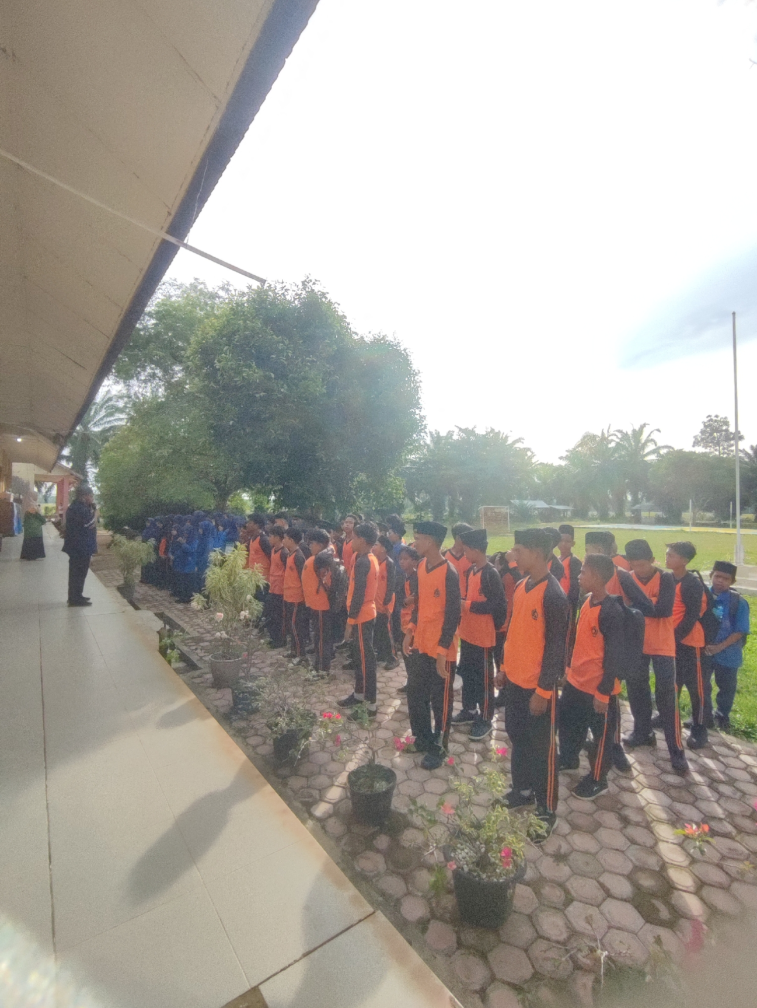A group of young people, likely students, are standing in formation on a paved outdoor area. The majority are wearing orange tops with black sleeves and trim, and black pants. Several are also wearing traditional headwear. They are lined up in neat rows, facing towards the left side of the frame. To the left, a person in a dark uniform, possibly a teacher or supervisor, stands facing the group. Behind the students, there is lush green vegetation, including trees and bushes, and a large, light-colored building with an overhang. The ground is a light-colored hexagonal paver. The sky is bright and hazy, suggesting it is daytime. The scene appears to be in a schoolyard or similar educational facility. The location is identified as Blangriek, Indonesia.