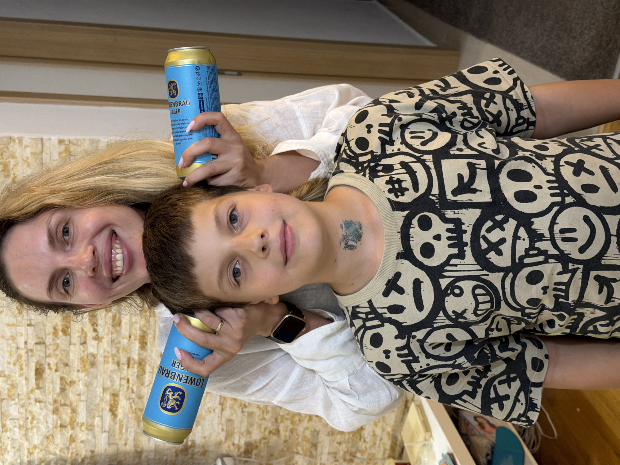 In Budva, Montenegro, a woman with blonde hair, wearing a white shirt, holds two beer cans, one over each side of a boy's head. The boy, wearing a black and beige t-shirt with a skull and smiley face pattern, looks towards the camera. He has a small, green, skull-shaped tattoo on his neck. The background consists of a light brown brick wall and a doorway with a carpet. The lighting is bright, indicating an indoor setting.