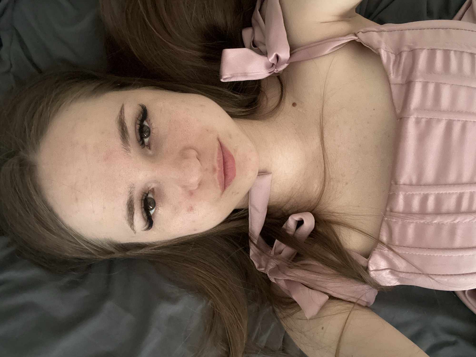 A woman is lying on a gray surface. She has long brown hair, fair skin, and is wearing a pink satin garment with ribbon ties. The location is Unknown, Unknown.