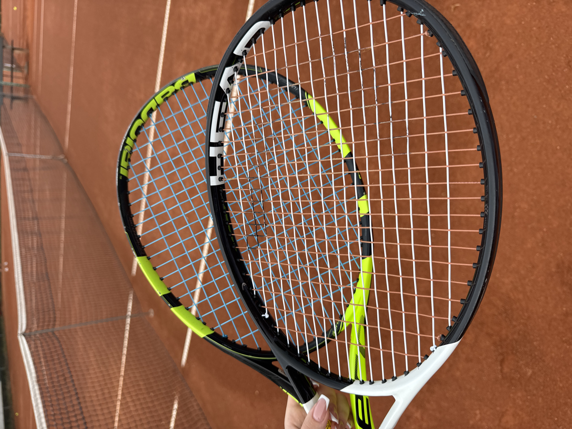 Two tennis racquets are held in a person's hand, one above the other, on a clay tennis court. The top racquet has blue strings, and the bottom racquet has copper-colored strings. The racquets are angled towards the camera, with the strings and frames clearly visible. The tennis court surface is a reddish-brown color, with white court lines visible in the background. A tennis net is also visible in the background, spanning across the court. The scene is lit with natural light, suggesting it is daytime. The location of Neubiberg, Germany is not evident from the image itself.