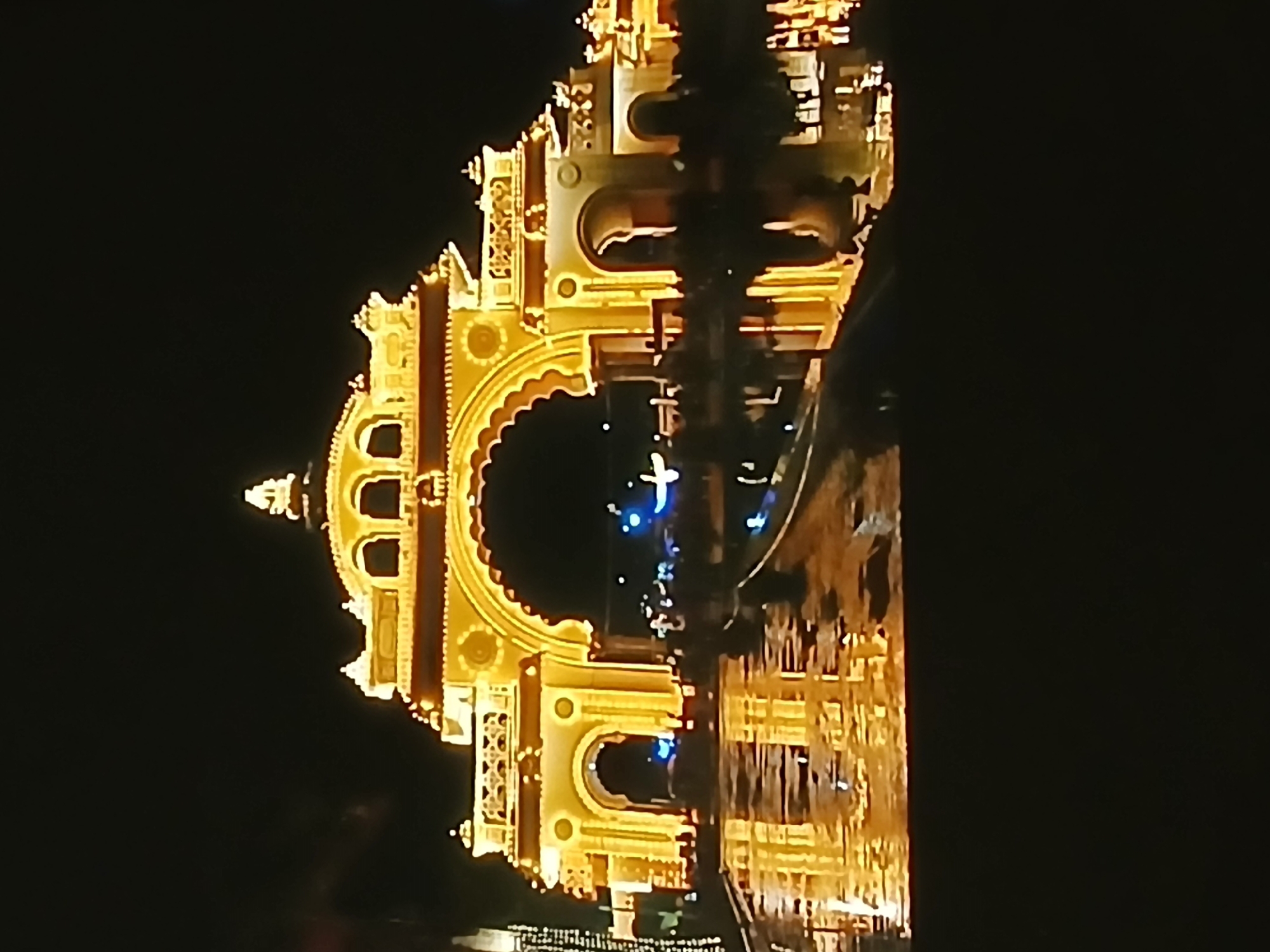Illuminated at night, the Mysore Palace is depicted with its intricate architectural details highlighted by warm yellow lighting. A body of water reflects the palace’s lights, creating a mirrored effect. The sky in the background is dark, emphasizing the brightness of the palace and its reflection. The palace is located in the city of Bengaluru, India.
