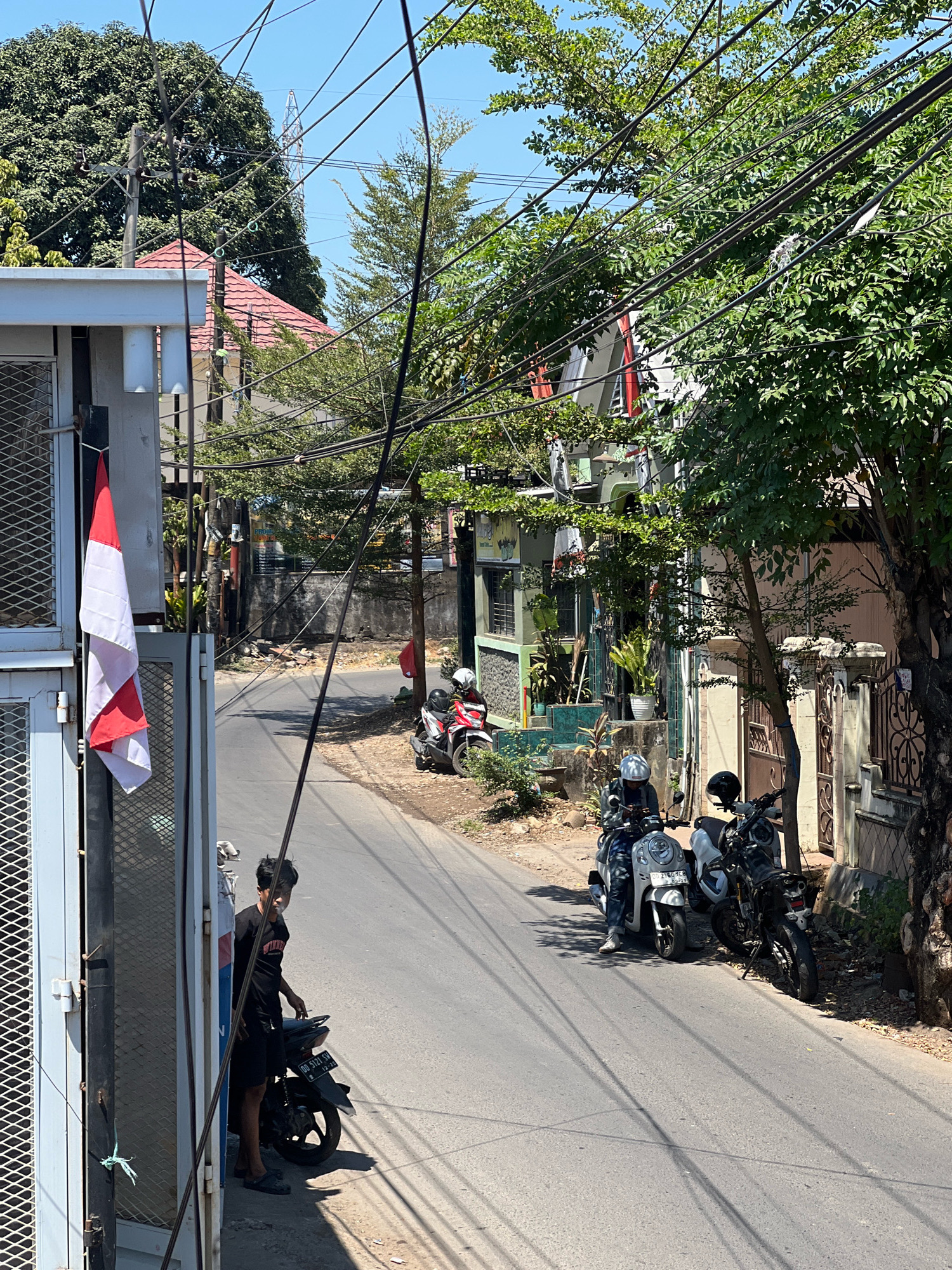 This image depicts a street scene in Makassar, Indonesia, under a clear blue sky. In the foreground, on the left, is a portion of a building with a grey fence and a white vertical metal structure. An Indonesian flag, red and white, is displayed prominently, hanging from a pole on this structure. A young man in a black t-shirt and black shorts stands near a parked motorcycle to the right of the flag.
The street itself is paved and slopes gently upwards and to the right. Several motorcycles are parked along the right side of the street. One red and white scooter with a rider wearing a white helmet is parked further up the street, with another white scooter parked close to it. Further down the street, two more motorcycles are visible.
The surroundings are lined with houses and vegetation. Trees with dense green foliage overhang the street, casting shadows. Buildings with red-tiled roofs and pastel-colored walls are visible. Overhead, a tangle of electrical wires crisscrosses the sky, supported by utility poles. The scene appears to be a residential or commercial street in a warm climate.