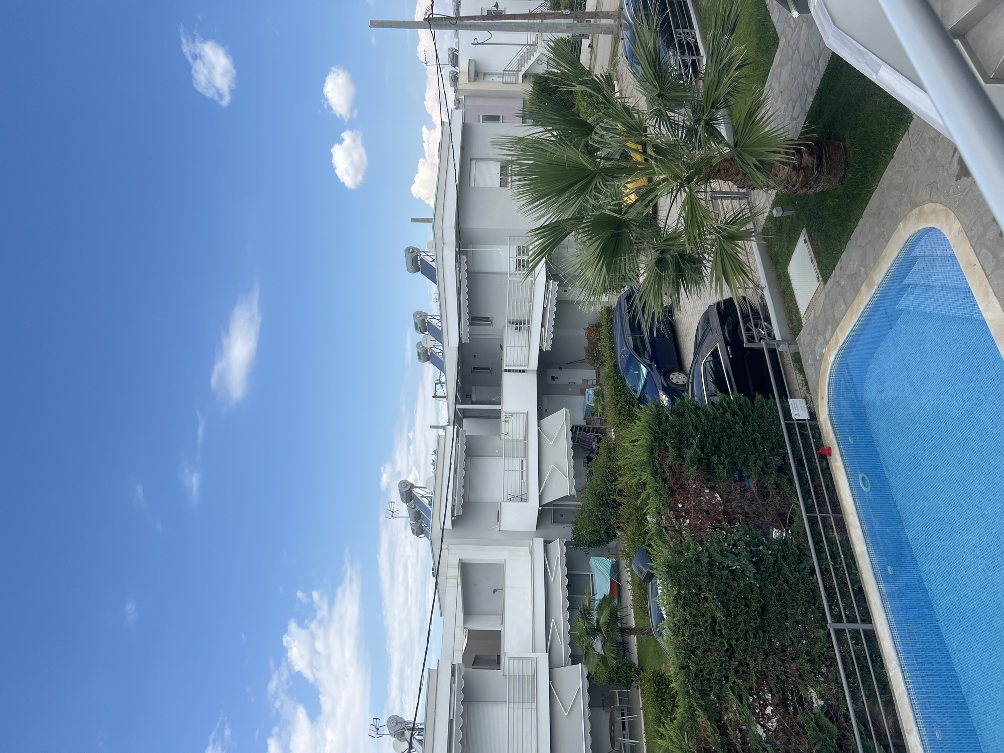 The image depicts a residential complex in Ofrynio Beach, Greece. In the foreground, a bright blue swimming pool with mosaic tiles is visible, bordered by a grey railing. Adjacent to the pool is a small lawn with a palm tree and a paved pathway. In the background, a row of modern, multi-story houses with white awnings and balconies can be seen. Several cars are parked in front of the houses, including a dark blue van, a black sedan, and a yellow car. The sky above is a clear blue with scattered white clouds. The scene appears tranquil, suggesting a residential or vacation setting.