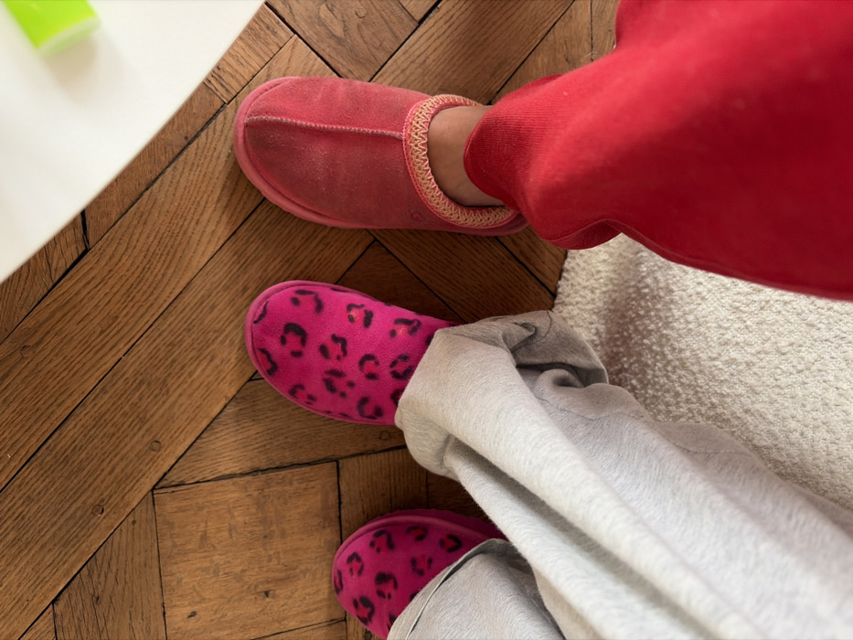 An overhead view captures the lower legs and slipper-clad feet of two individuals in an indoor setting. The floor primarily consists of light to medium brown herringbone parquet, indicative of classic Parisian apartment interiors. A segment of a light-colored, textured rug is visible in the foreground. In the upper right corner, a portion of a white surface, possibly a table, is visible with a blurred green object resting upon it. The individual on the left wears grey trousers and bright pink slippers adorned with a black leopard print. The individual on the right wears red trousers and a single red, plush slipper with a light-colored braided trim around the ankle opening. Both subjects appear static.