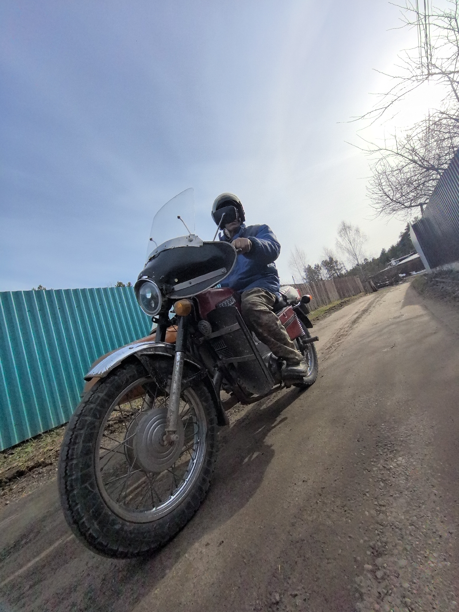 A man wearing a helmet and blue jacket is riding a vintage motorcycle on a dirt road in Noviy, Russia. The motorcycle has a windshield, and a fence and trees line the road.